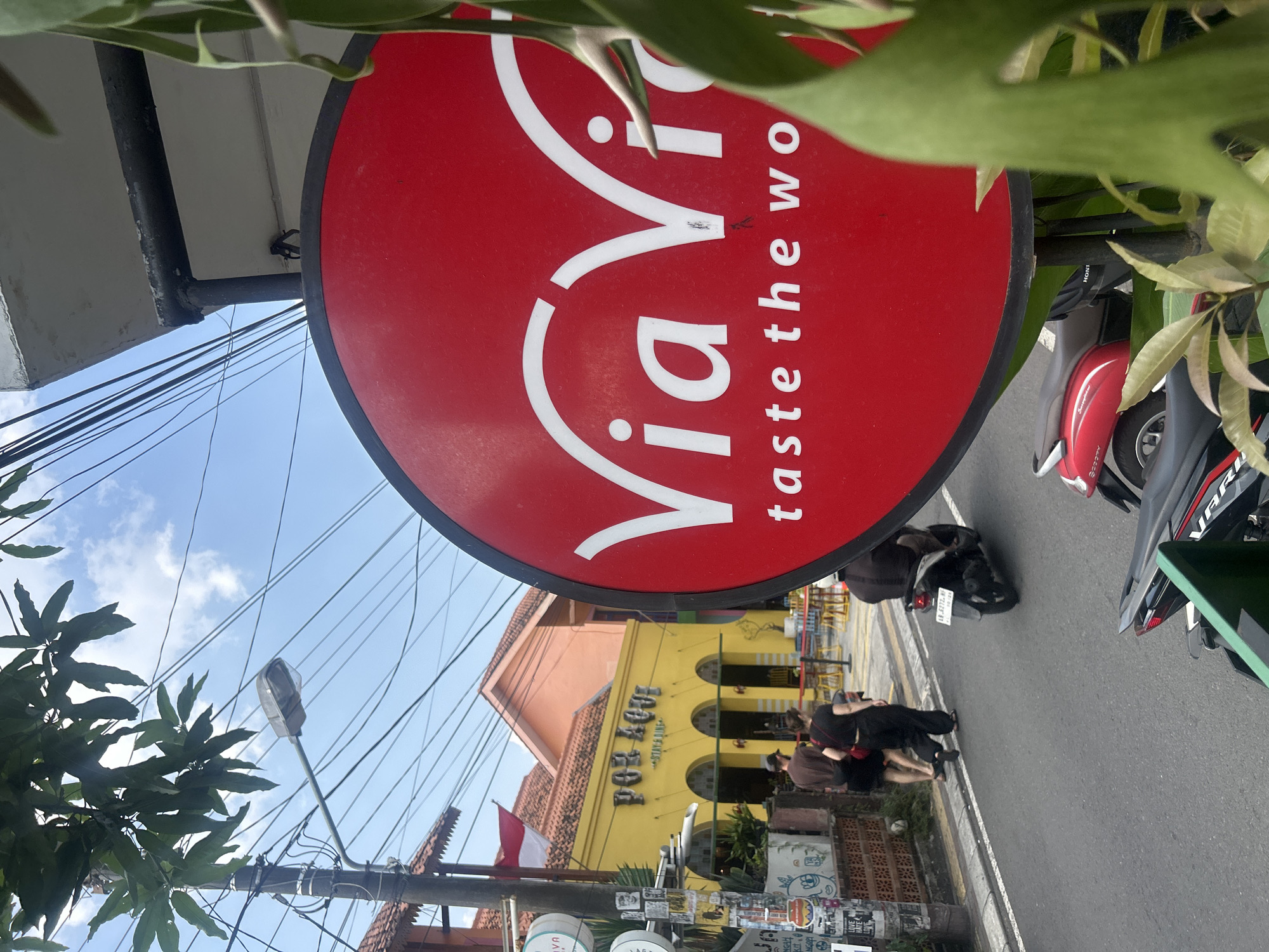 The image depicts a street scene in Yogyakarta, Indonesia, focusing on a bright red circular sign with white text. The sign reads "Via Via" at the top, with the tagline "taste the wo" visible below. To the left of the sign, a yellow building with arched windows and the text "POR AQUL" and "STAY & DINE" is visible. Two individuals, a man and a woman, are walking on the sidewalk in front of the yellow building. A man is riding a scooter in the street, heading away from the viewer. In the foreground, a portion of a green plant is obscuring the lower right side of the red sign. Overhead, a clear blue sky is visible with numerous electrical wires. The overall scene suggests a commercial area with a lively atmosphere, typical of a tourist destination in Yogyakarta.
