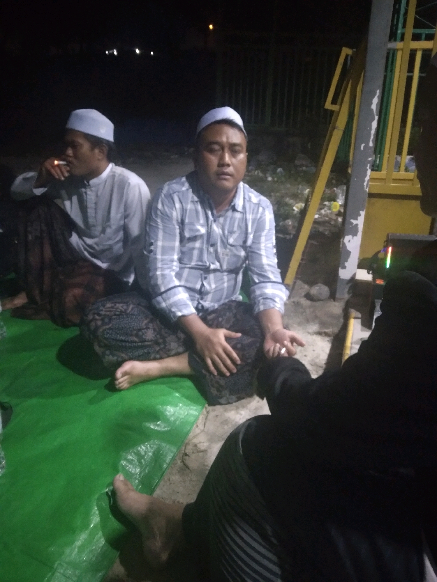 Three individuals are seated on a green tarp at night, possibly in an outdoor area of Batam, Indonesia, given the context. The person on the left, wearing a white peci and a light-colored, long-sleeved shirt, is smoking a cigarette. They are also wearing a patterned sarong. To their right, another person, also wearing a white peci and a checkered shirt, is seated with their hands in their lap. This person is also wearing a patterned sarong. A third individual, mostly obscured, is wearing dark clothing and is seated on the right side of the frame, facing away from the camera. In the background, there is a metal structure with yellow and green accents, and further beyond, some indistinct lights are visible against a dark sky, suggesting an urban or semi-urban setting. The ground appears to be a mix of dirt and concrete.