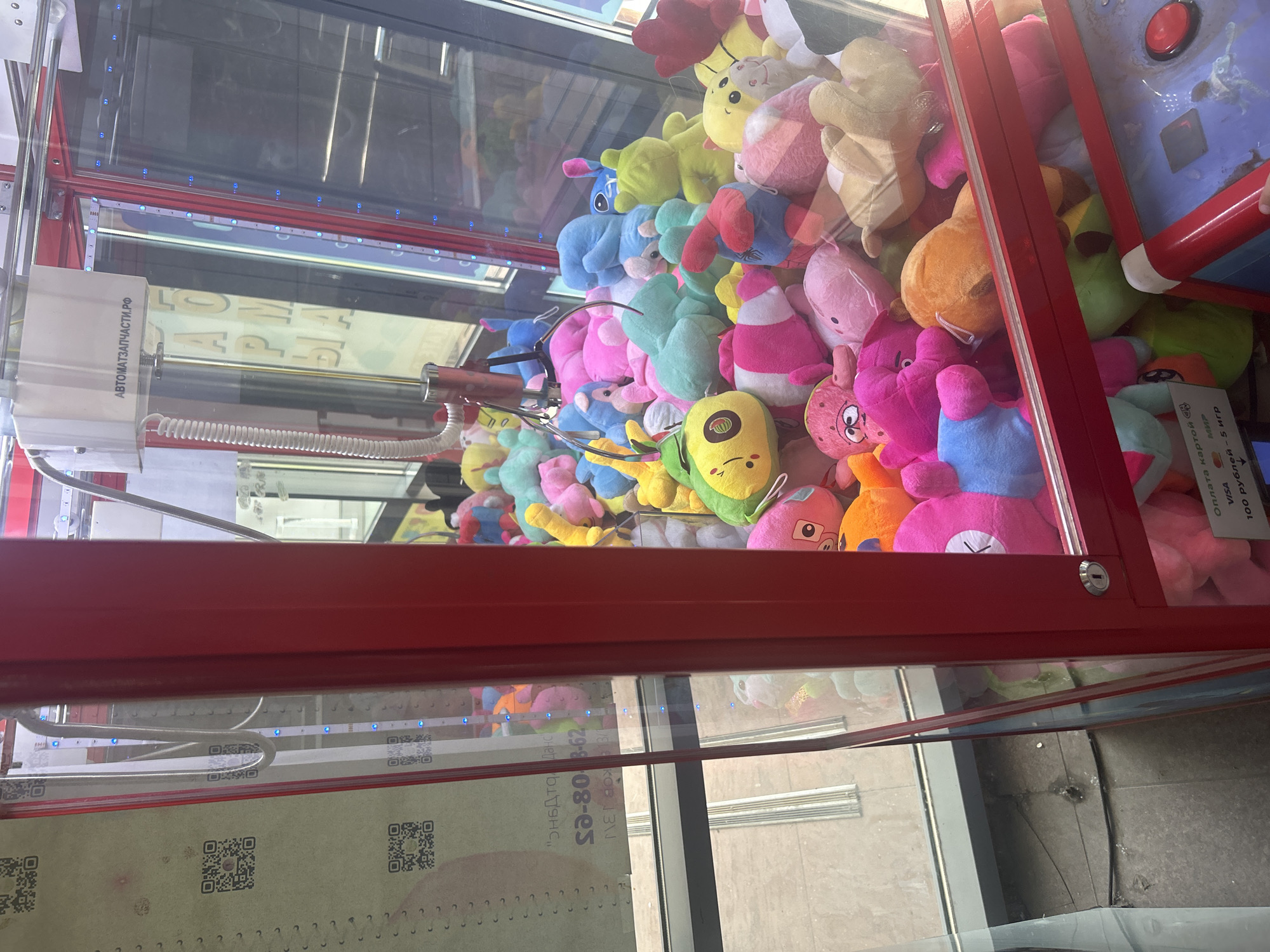 This media file is a close-up, slightly angled shot of the interior of a brightly lit claw machine filled with a variety of plush toys. The machine's exterior is a vibrant red metal frame with glass panels. Inside, a dense assortment of colorful stuffed animals, predominantly pink, blue, yellow, and green, are packed together. Some of the toys appear to be animal-shaped, with ears, snouts, and bright, cartoonish eyes.
The background within the machine is somewhat obscured by the toys, but reflections on the glass suggest an indoor environment, possibly a mall or arcade. Visible in the lower section of the image are QR codes and some text printed on paper. The text, partially legible through the glass, includes "Оплата картой" (Payment by card), "VISA", "МИР", and "100 рублей - 5 игр" (100 rubles - 5 games), indicating this is a coin-operated redemption game. Another visible piece of text on the machine's components reads "АВТОМАТЗАПЧАСТИ.РФ", suggesting it's related to automated parts.
The lighting appears to be artificial and bright, illuminating the toys and highlighting the reflective surfaces of the glass. No people are visible in the frame, but the presence of the claw machine implies it is in a public space where people would play. The overall scene depicts a typical arcade game setting, focused on the colorful and inviting display of prizes within the machine. The exact time of day and weather are not discernible from the image. The context provided indicates the location is Saint Petersburg, Russia.