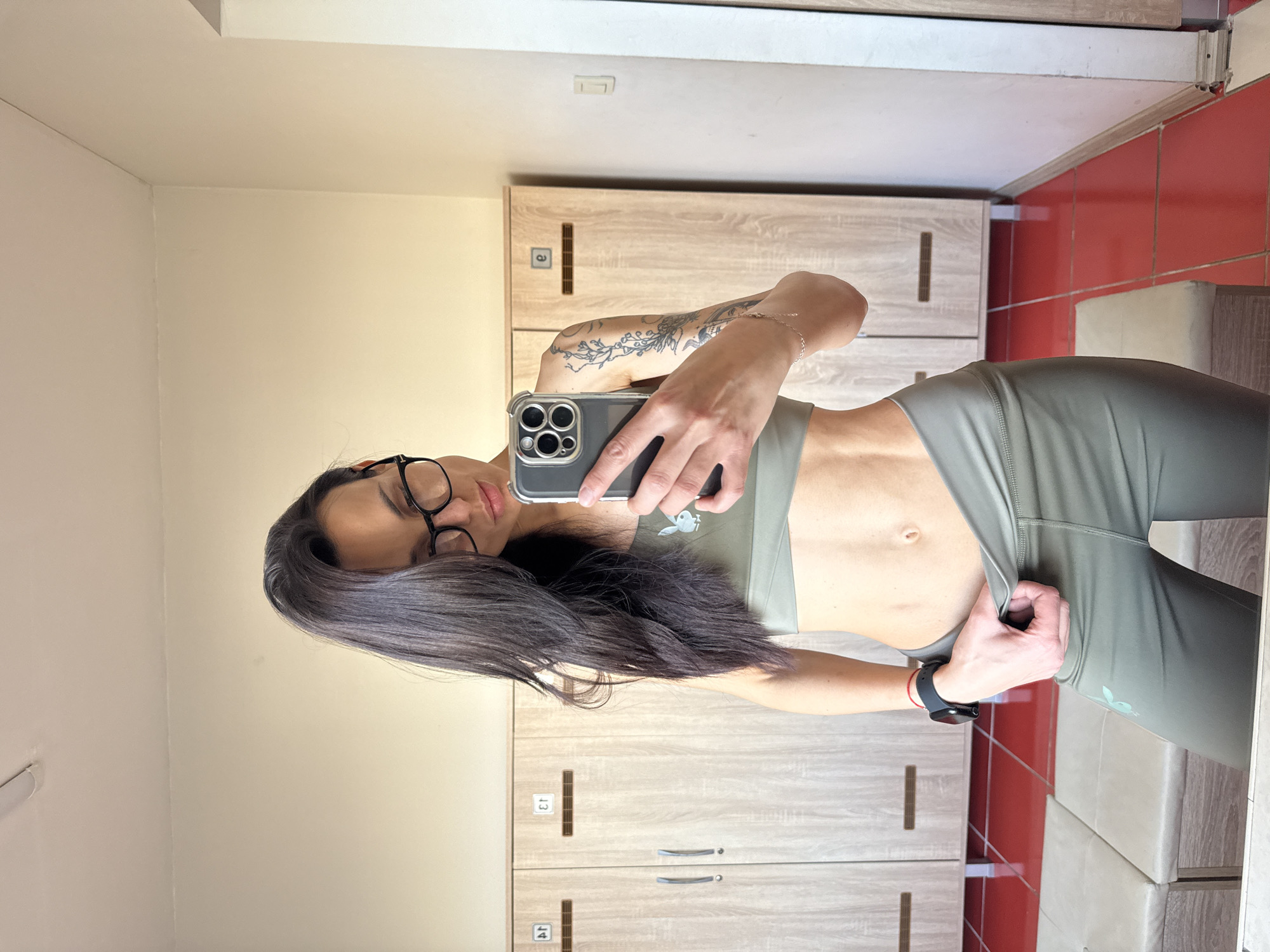 A woman is taking a selfie in a locker room. She is wearing a matching sports bra and leggings set. She has long dark hair and is wearing glasses. The background includes a row of lockers, red tiled walls, and a white wall. The lockers have numbered tags and horizontal handles. The woman is holding a smartphone. The location is near the city of Pleven, Bulgaria.