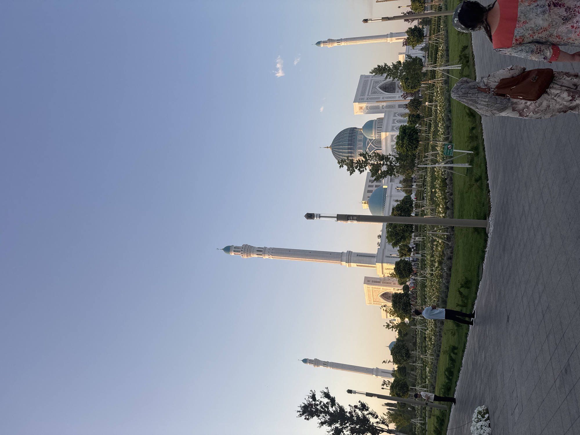 In the city of Khoja Ismail, Uzbekistan, a grand mosque complex is pictured during the golden hour of sunset. The foreground features a paved walkway where two women are walking away from the camera, their backs to the viewer. The woman on the left wears a patterned headscarf and a long, flowing dress, carrying a brown purse over her shoulder. The woman on the right has a dark headscarf and a colorful dress. Further in the background, two men are standing in a manicured garden area. One man in a white shirt and dark pants stands near the edge of the walkway, while another man in a light blue shirt and dark pants stands further back.
The mosque complex itself is a dominant feature. It is a large, white structure with multiple domes and tall, slender minarets. The main dome is a striking turquoise color, and several smaller turquoise domes are also visible. The minarets are tall and white, topped with turquoise accents. The architecture suggests a classic Islamic style. The surrounding area is landscaped with a variety of trees and manicured bushes, many of which are staked. The sky is a clear, soft blue, transitioning to a warmer hue near the horizon, indicating the setting sun. The overall scene conveys a sense of peace and grandeur.