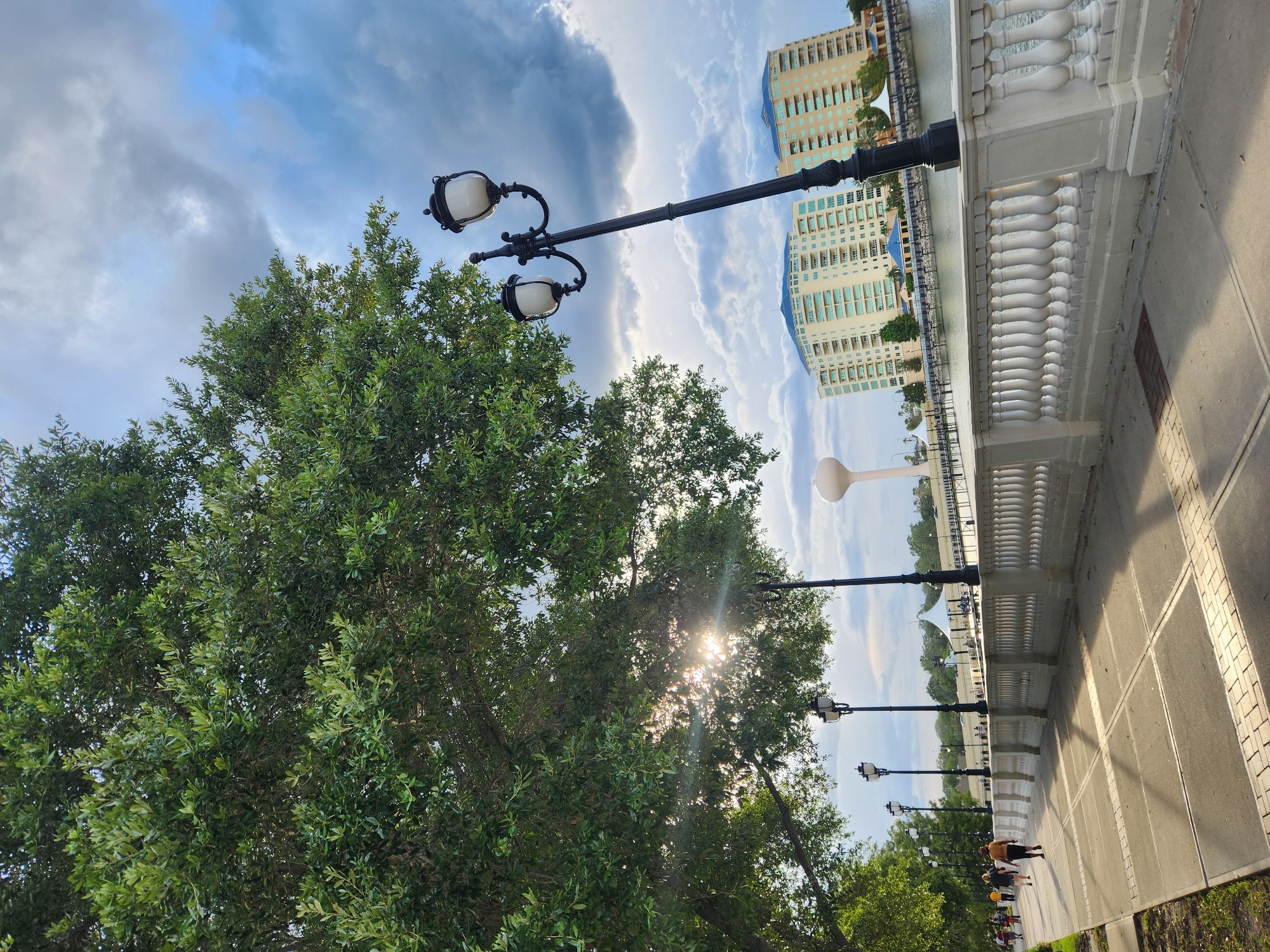 The image depicts a pedestrian walkway alongside a body of water, likely a lake or river, in Altamonte Springs, United States of America. A large, leafy tree dominates the left side of the frame, with sunlight filtering through its branches. Along the walkway, several ornate streetlights stand tall. In the background, two multi-story buildings with blue roofs are visible, situated across the water. A white water tower is also present in the distance. People are seen walking along the path, and a white, ornate railing lines the edge of the walkway overlooking the water. The sky is partly cloudy with a mix of sunlight and dark clouds.