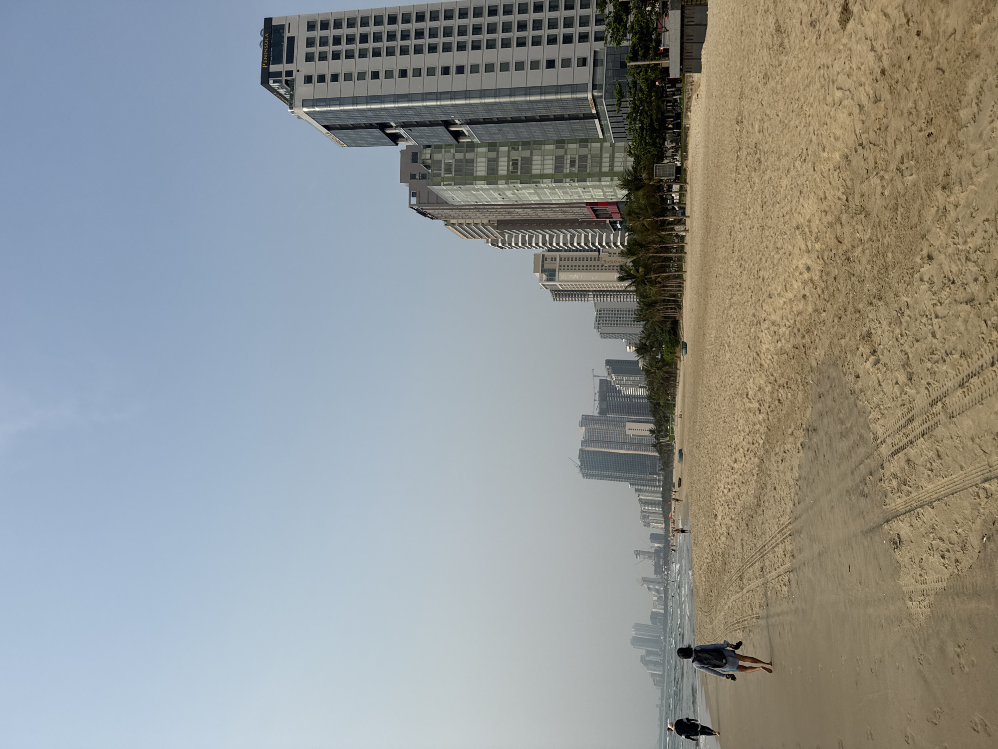 The image shows a beach scene in Đà Nẵng, Vietnam. The shore has tire tracks. Several high-rise buildings line the coast, including a building labeled "PENINSULA." Two individuals are seen walking along the beach. The sky is hazy.