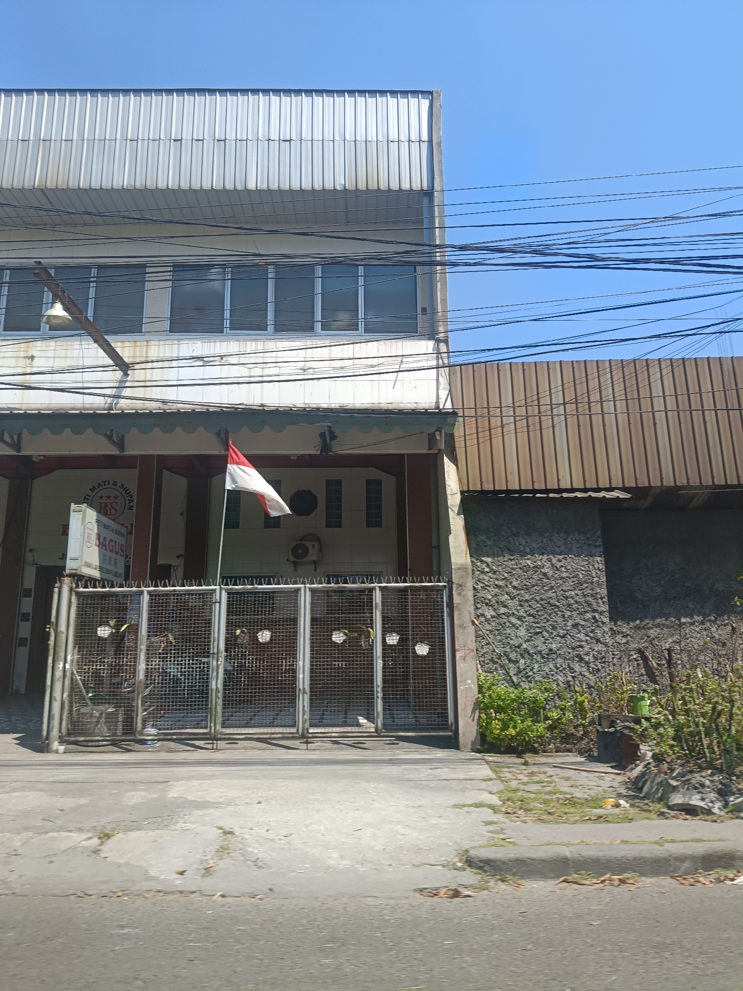 The image captures a view of a building in Surabaya, Indonesia, under a clear blue sky. The building has a corrugated metal roof and a light-colored facade. There are several windows with a reflective glaze on the upper floor. Below, there's an awning with decorative scalloped trim. A gate made of metal bars is visible in the foreground, with a small Indonesian flag displayed on a pole. Behind the gate, there's an entrance to the building, with a sign that appears to be in Indonesian. An air conditioning unit is mounted on the wall near the entrance. To the right of the main building, there's another structure with a corrugated metal roof and a textured, gray stone-like wall. Some vegetation and dry leaves are scattered around the base of this structure and the sidewalk. Electrical wires are strung across the top of the scene, indicating an urban environment. The street in front of the building is paved with asphalt and shows some wear and tear.