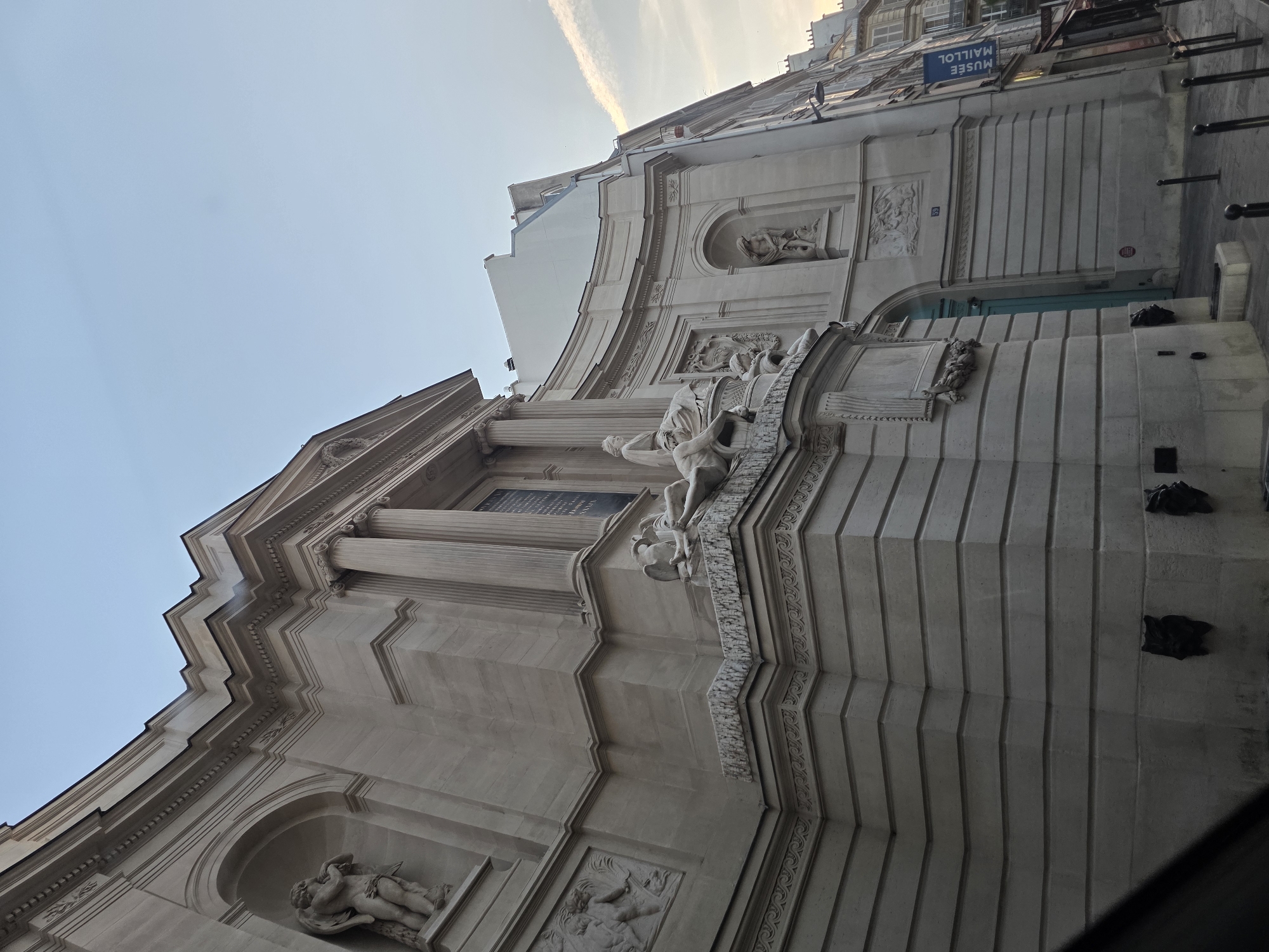 The image shows the exterior of a building in Paris, France. The building is made of light-colored stone and features classical architectural elements such as columns, arches, and sculptures. In the foreground, a portion of a dark object, possibly a vehicle, is visible. The building has intricate carvings and statues adorning its facade, including what appears to be a depiction of cherubs and a reclining figure. A blue door is visible, and a sign that reads "MUSÉE MAILLOL" is hanging on the side of the building, indicating it is the Maillol Museum. The street in front of the museum is paved, and there are lampposts and bollards lining the sidewalk. The sky is a pale blue with a wispy cloud. The overall scene suggests a street view of a museum in Paris.