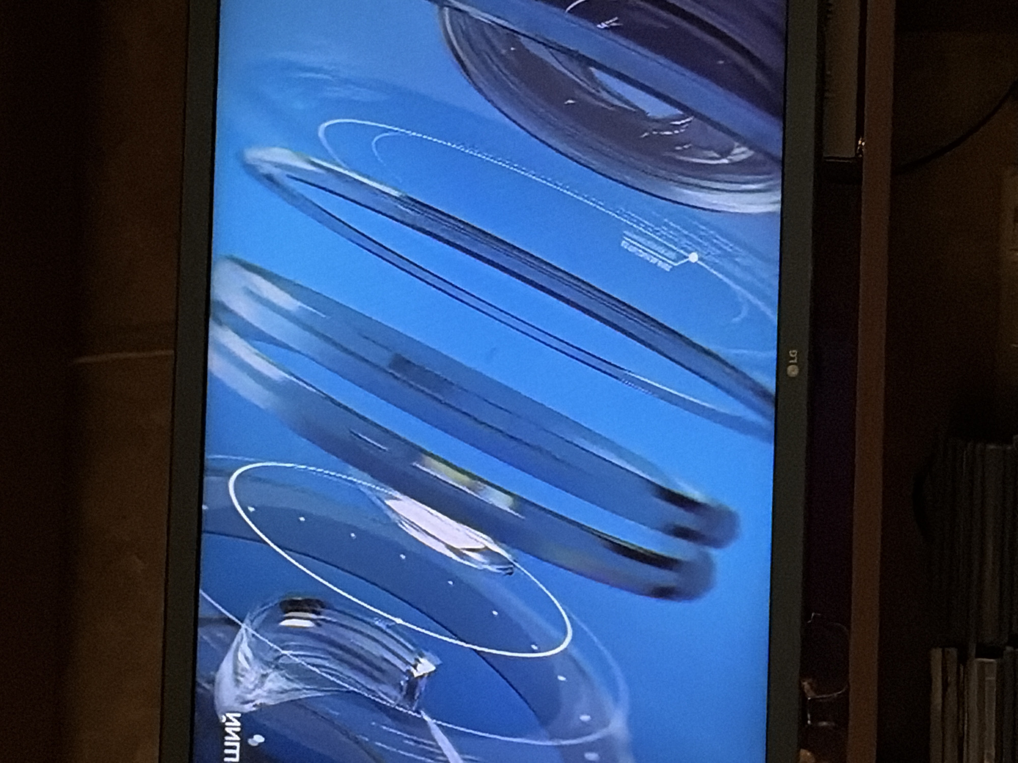 The image shows a close-up view of a television screen displaying a news program or similar broadcast. The screen is predominantly blue with abstract, metallic-looking graphic elements that appear to be swirling or moving. In the lower left corner of the screen, the LG logo is visible. The television itself is dark grey and appears to be sitting on a shelf or furniture. Below the television, a stack of what appear to be DVDs or CDs is visible. The background outside the frame of the television is dark and appears to be a wall or room interior, with some wooden furniture. There are no people or discernible city elements visible in the image.