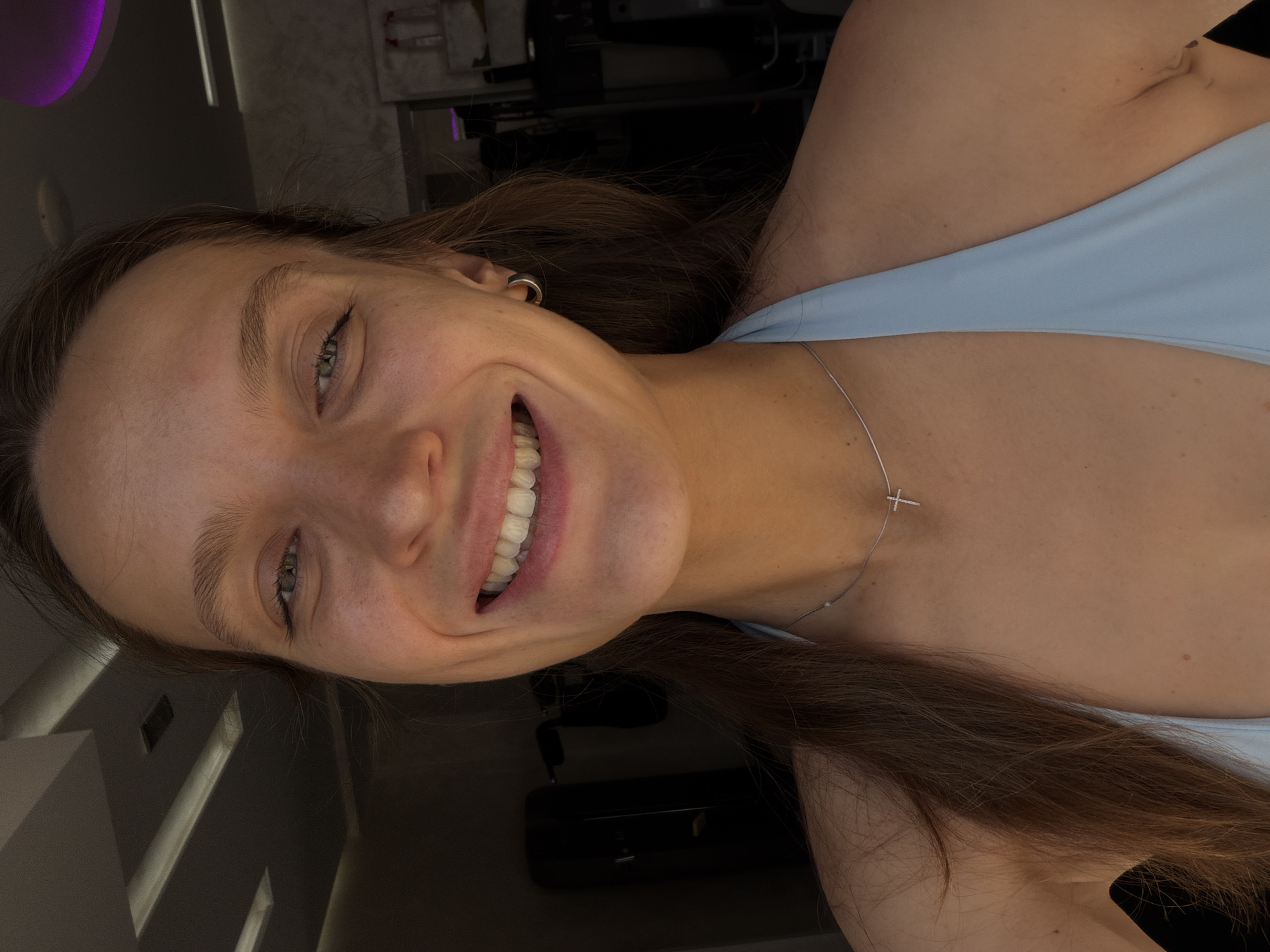 A young woman with light brown hair smiles directly at the camera. She is wearing a light blue halter top and a thin silver necklace with a cross pendant. She has a hoop earring in her right ear. The background appears to be indoors, possibly a gym, with exercise equipment visible. The lighting suggests it is daytime. The specific location in Budva, Montenegro, is not discernible from the image.