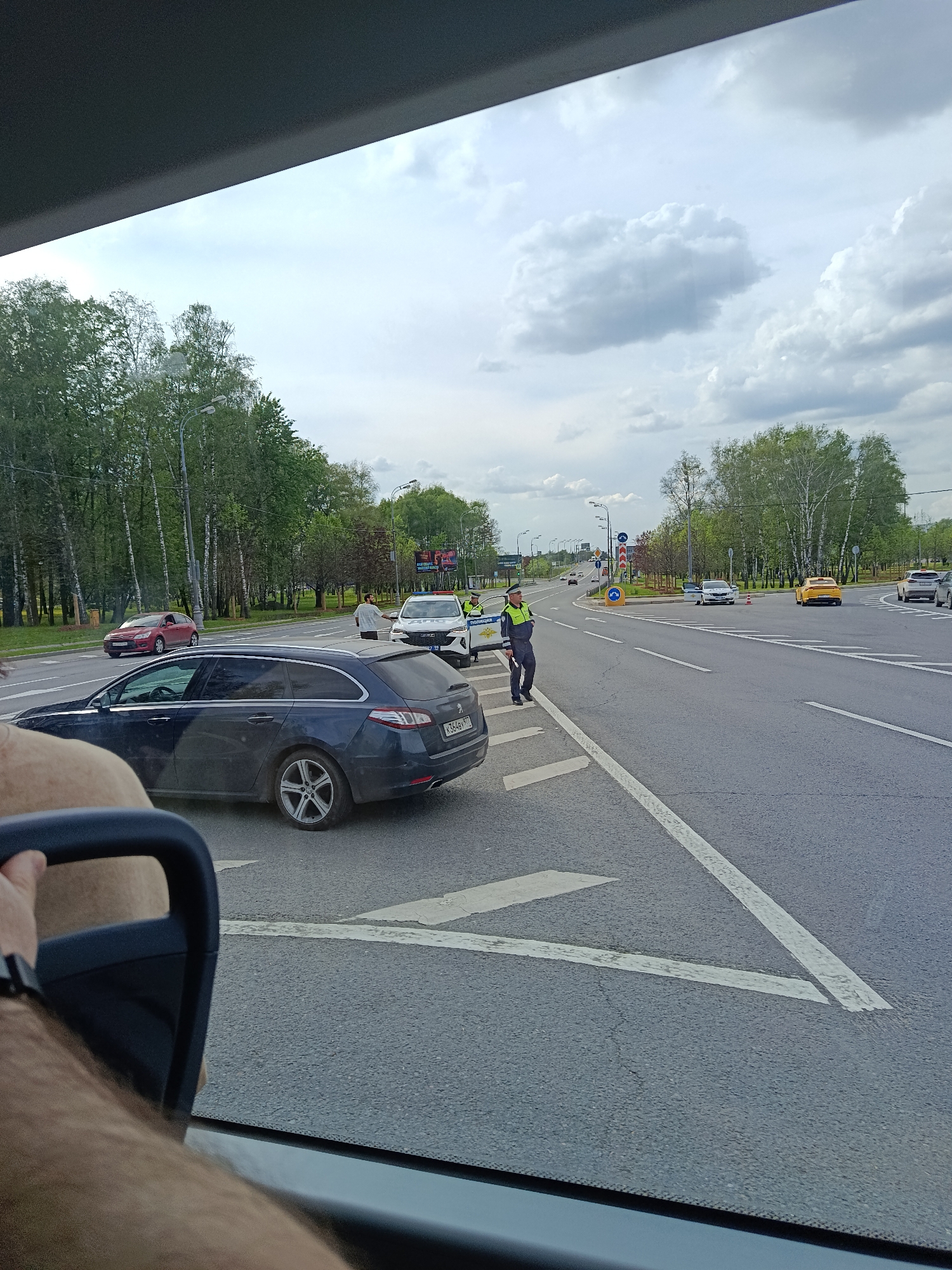 A photograph taken from inside a vehicle shows a traffic stop on a road in Moscow, Russia. A dark blue Peugeot station wagon with license plate number K364BV 777 is pulled over on the road's shoulder. A police officer wearing a neon green and blue uniform is standing beside the Peugeot, interacting with the driver. Another person in a grey shirt stands near the police car. A white police vehicle is positioned behind the Peugeot. The intersection ahead includes a roundabout. Other cars can be seen driving on the road. Trees line the side of the road and there are clouds in the sky.
