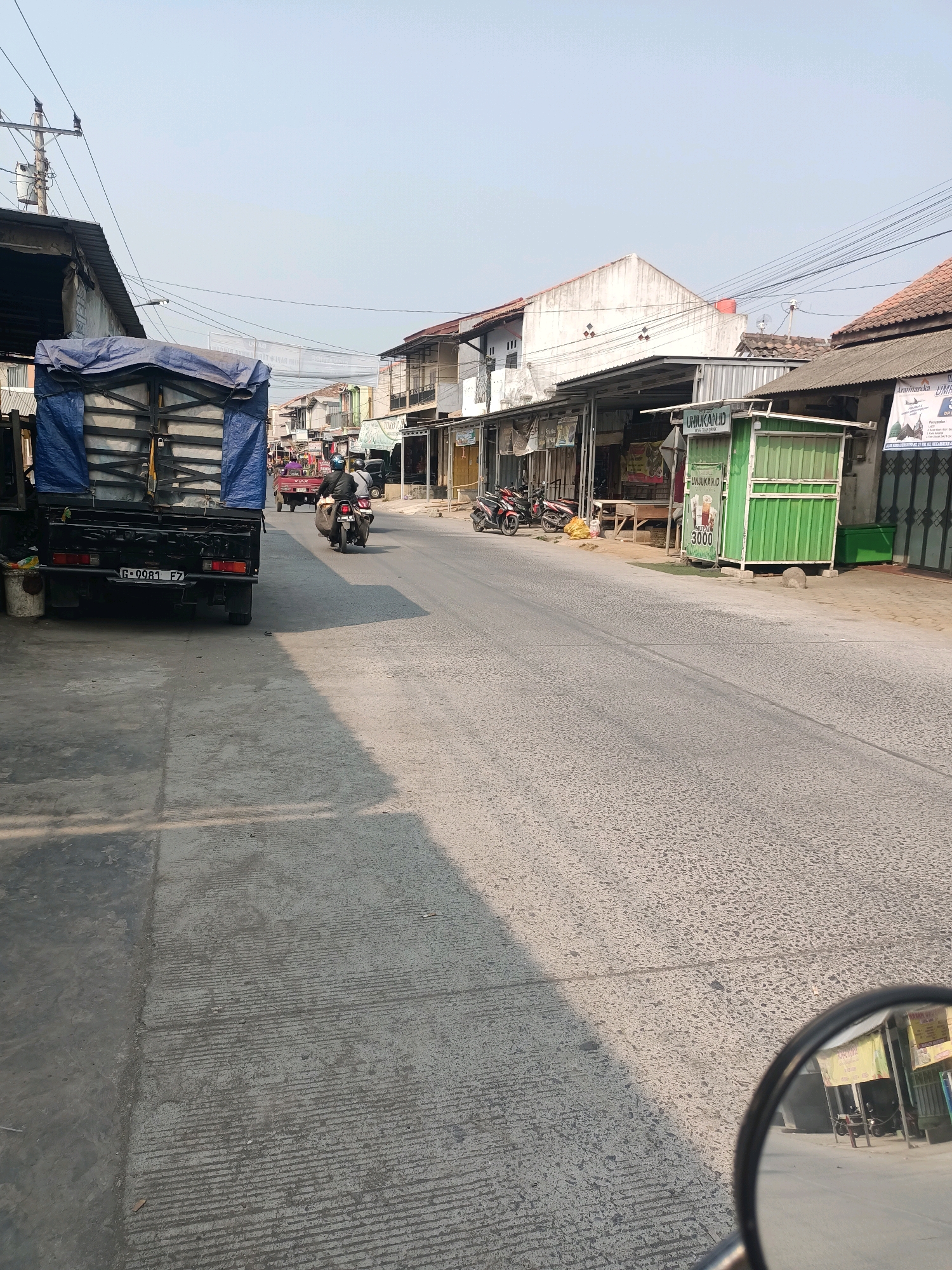The image captures a street scene in Tembok Banjaran, Indonesia, under a clear sky. In the foreground, a portion of a motorcycle's side mirror is visible, reflecting the street ahead. The road is paved with a textured asphalt surface. To the left of the road, a parked black truck with a blue tarp covering its cargo is positioned on a concrete area. The truck has a license plate reading "G-9981 FZ". Beyond the truck, a row of low-rise buildings with various storefronts lines the right side of the street. One prominent green stall with the text "UNJUKAN.ID" and a price "3000" is visible. Several motorcycles are parked in front of the storefronts. Further down the street, a few people are riding motorcycles. The buildings exhibit traditional Indonesian architecture with exposed roof tiles and varying facade designs. Power lines and utility poles are present along the street. The overall impression is a bustling commercial street in a residential area.