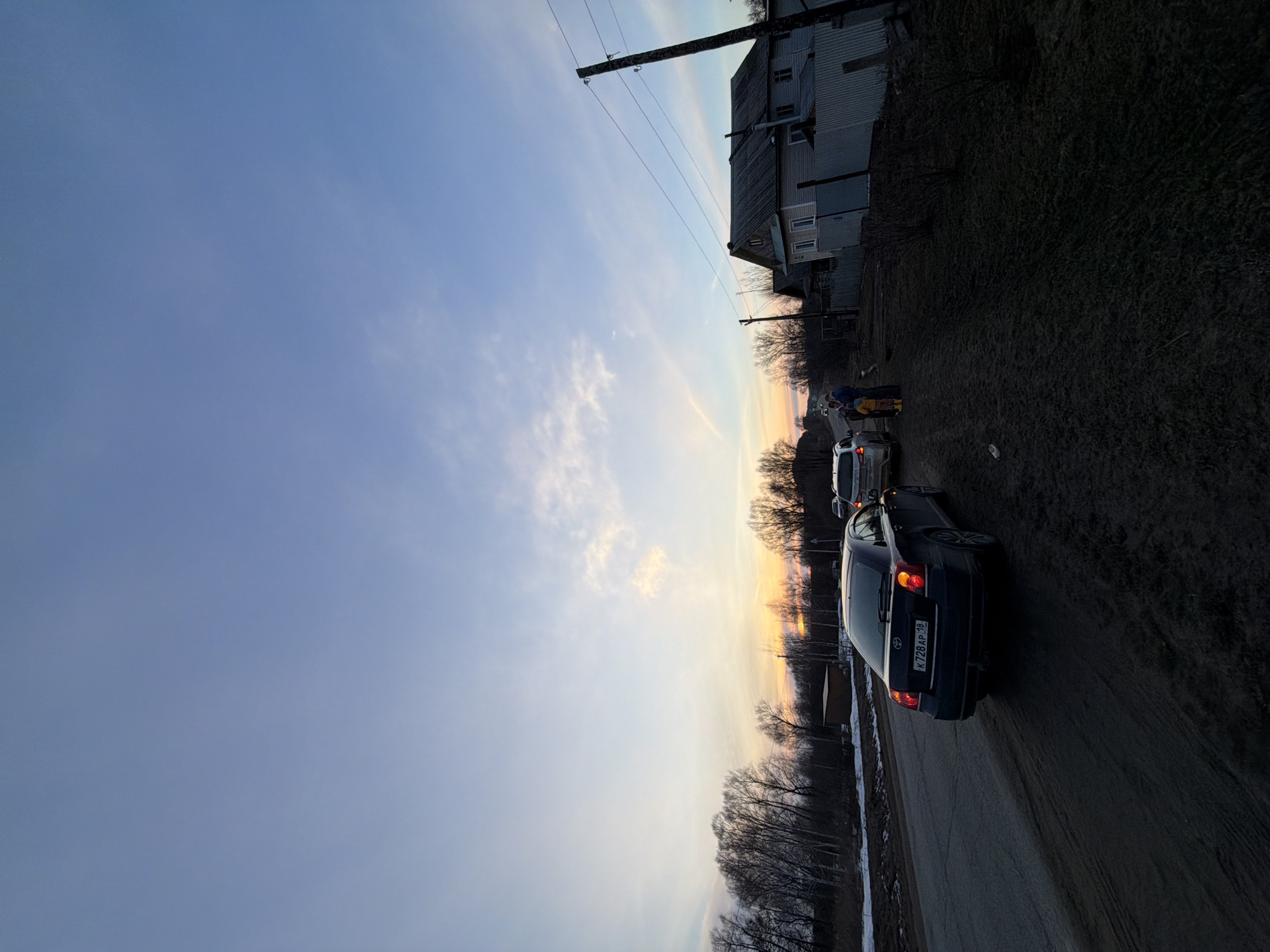 The image depicts a street in Дулесово, Russia, at dusk. The sky is a gradient of blue and orange, indicating a sunset. A grey, two-story building stands on the right side of the frame, adjacent to a grassy area. Utility poles with wires run alongside the building. Several cars are positioned on the road, with a dark-colored car being the closest to the viewer. The license plate on the back of this car shows "К 728 AP 10". Two people are standing next to one of the vehicles further down the road. Bare trees are visible in the background.