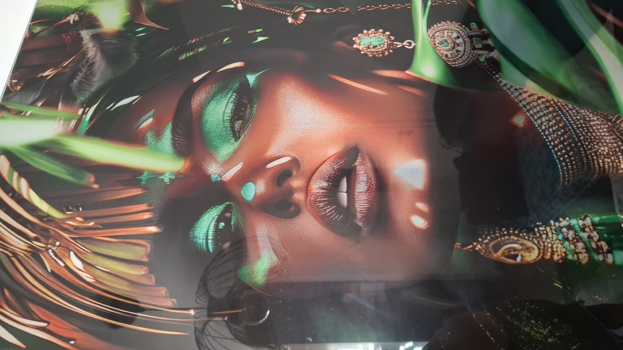 The image is a close-up, studio portrait of a woman with dramatic makeup. She has vibrant green eyeshadow that extends to her eyebrows and under her eyes, and a turquoise gem on her forehead. Her lips are dark and glossy, and her skin has a warm, bronzed tone with highlights and shadows. She is adorned with intricate gold jewelry, including dangling earrings with green stones and a layered necklace with similar embellishments. The background is dark and out of focus, with hints of green foliage and possibly feathers. There are some reflective or transparent elements in the foreground, obscuring parts of the image.
The image is a photograph, likely of a painting or print, displayed within a frame. The overall aesthetic suggests a stylized and artistic representation, possibly for fashion or editorial purposes.
There is no discernible information in the image to place it in the city of Buchen (Odenwald), Germany.