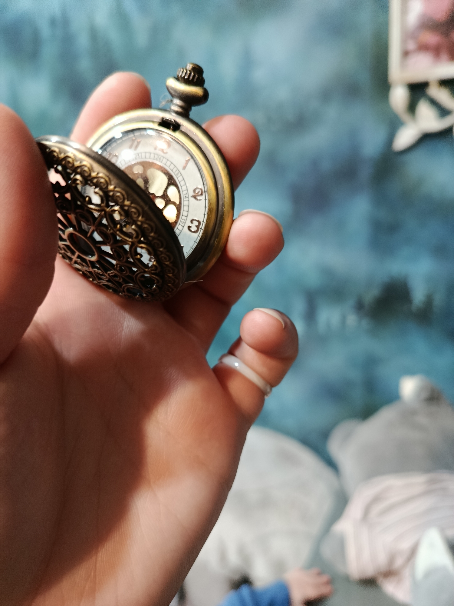 A person's hand is holding an open, ornate pocket watch with a brass-colored casing and a filigree cover. The watch face is visible, showing Roman numerals and intricate inner workings. The background is a blurry blue wall with a textured pattern. In the lower right corner, parts of what appear to be stuffed animals and a person's arm are visible, suggesting an indoor domestic setting. The image does not contain any information about the city of Apatity, Russia.