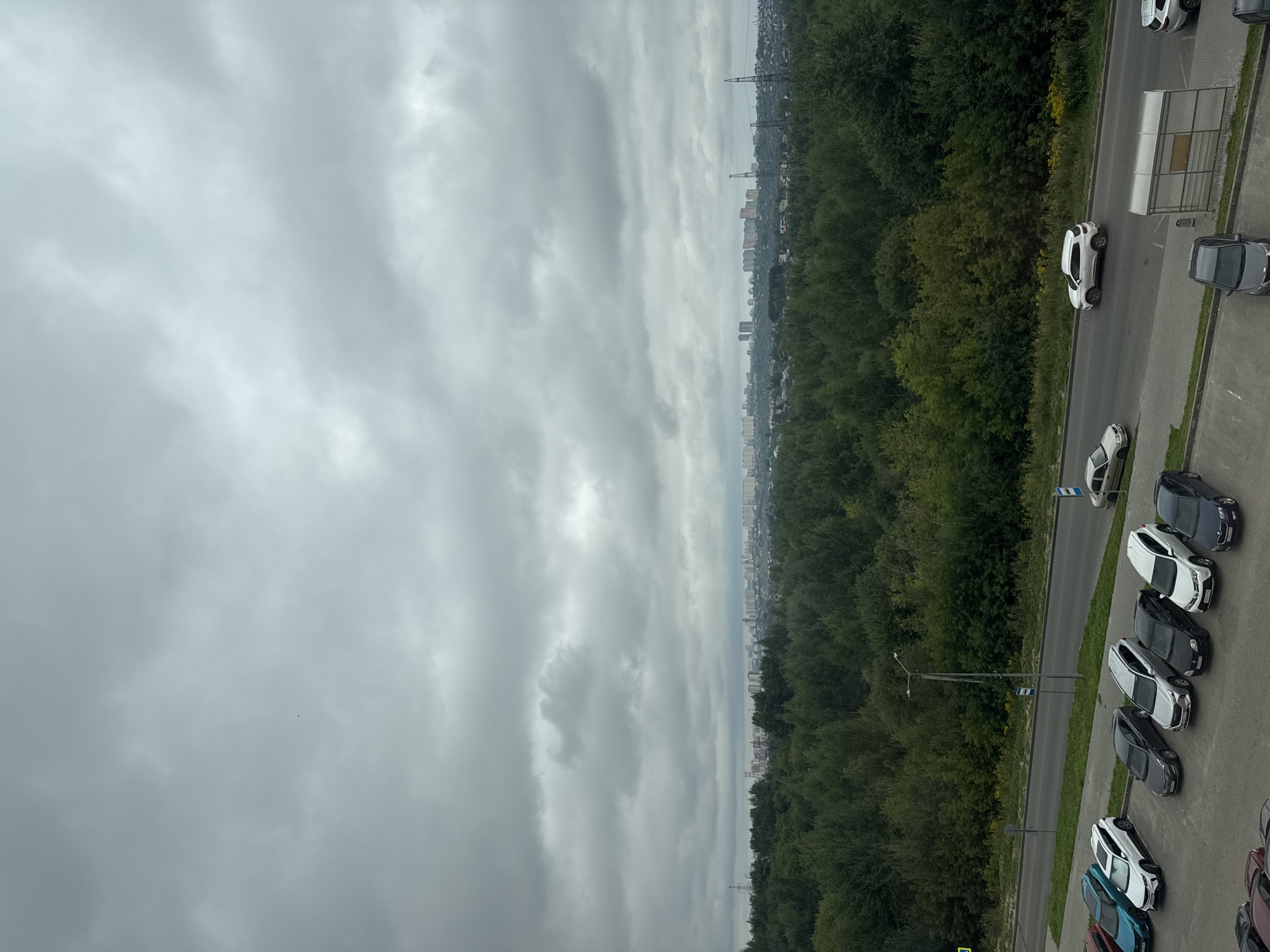 The image depicts a cloudy, overcast sky over a semi-urban landscape. In the foreground, a parking lot with several cars is visible. Cars of various colors, including white, black, silver, and blue, are parked in designated spots. A road runs parallel to the parking lot, with a few cars driving on it, including a white SUV and a silver sedan. Beyond the road is a dense forest of green trees, which stretches towards a distant city skyline. The city features numerous buildings, including apartment blocks and taller structures, silhouetted against the muted sky. Electrical pylons are also visible in the middle ground. The overall impression is one of a subdued, possibly cool, day.