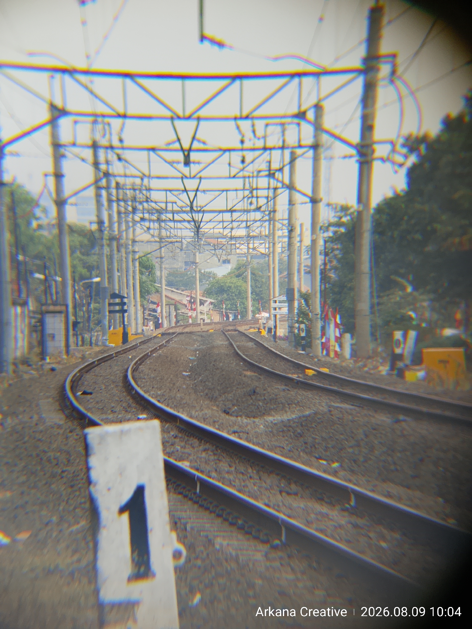 An outdoor, eye-level, medium shot captures a pair of train tracks curving away from the viewer into the distance, framed by a network of overhead power lines and poles. The tracks are made of gravel and metal rails, and on the left side of the image, a concrete marker with the number "1" painted in dark blue is partially visible in the foreground. The scene is hazy, suggesting a bright, possibly overcast or smoggy day. The background shows trees and some buildings, including what appears to be a structure with red and white flags, hinting at a location in a semi-urban or industrial area. The timestamp "Arkana Creative | 2026.08.09 10:04" is overlaid on the bottom right of the image, indicating the date and time of capture. The image has a chromatic aberration effect, giving a colorful halo around objects. The specific location in Bekasi, Indonesia, is not explicitly shown but can be inferred from the timestamp data.