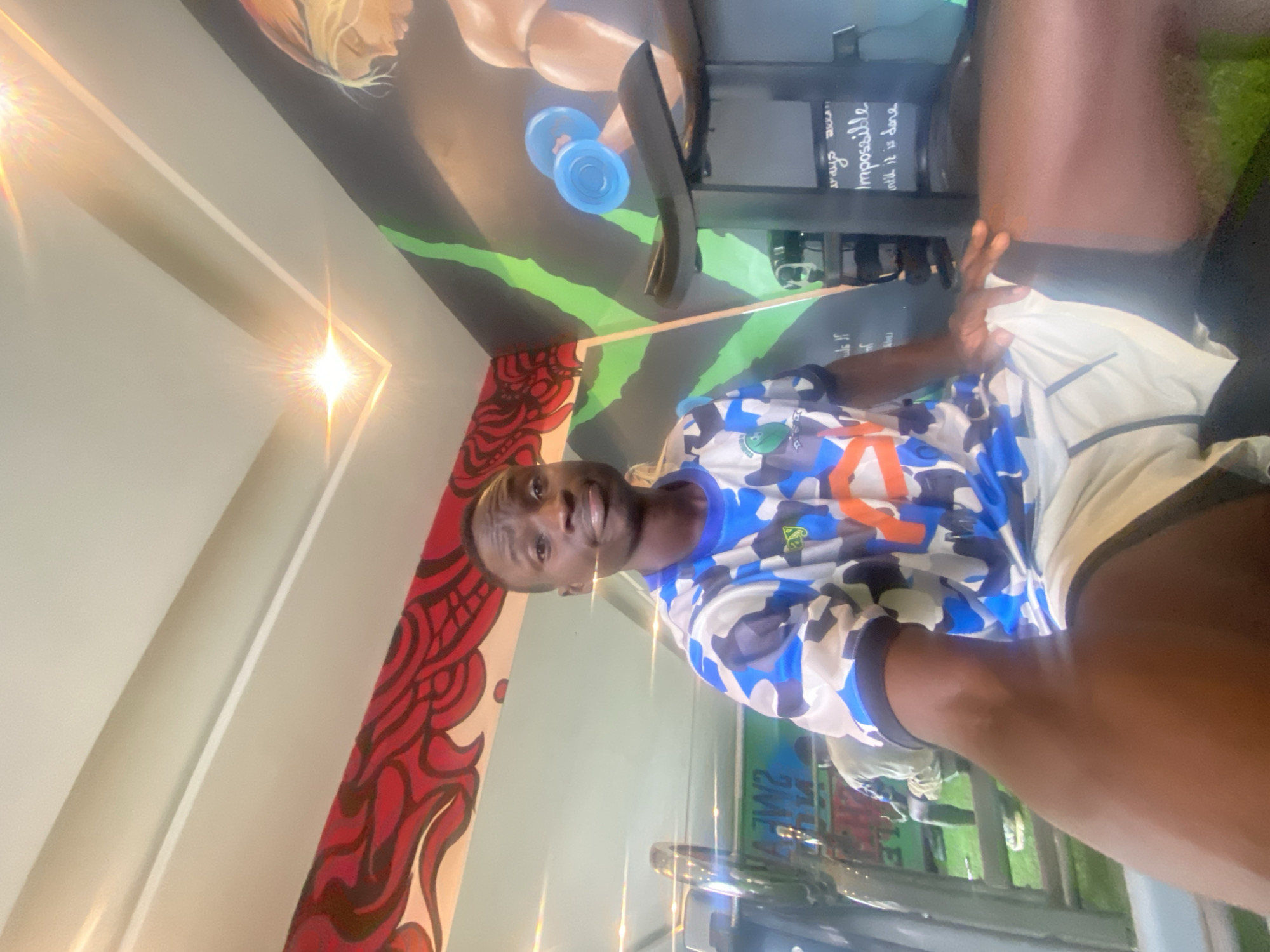 A man is seated, facing forward, in a gym. He is wearing a blue, white, and orange camouflage t-shirt and white shorts. He is holding his right arm in the foreground, resting his hand on his leg. Behind him, to the left, is a treadmill, and to the right is a wall mural depicting a muscular arm lifting dumbbells. The gym is illuminated by overhead lights, one of which is prominently visible on the ceiling. The background features a red and black pattern on the wall above a green stripe. The overall setting appears to be an indoor fitness facility. The location in the city of Barden Yana, Nigeria, is not determinable from the visual information provided in the image.