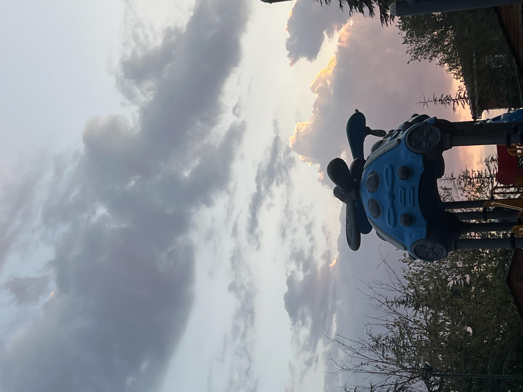A blue, plastic playground structure resembling a stylized beetle-car is positioned centrally in the frame, partially obscuring a sunset sky. The structure features two large, black, wing-like elements on top, and round, black protrusions resembling wheels on its sides. The sky above is filled with clouds, some dark gray and others illuminated by the setting sun with warm, golden and pink hues. Trees, including pine trees, are visible in the background, framing the scene and extending from the bottom edges of the image upwards. The overall setting suggests an outdoor public space, possibly a park or a playground in Keçiören, Turkey, during twilight.
