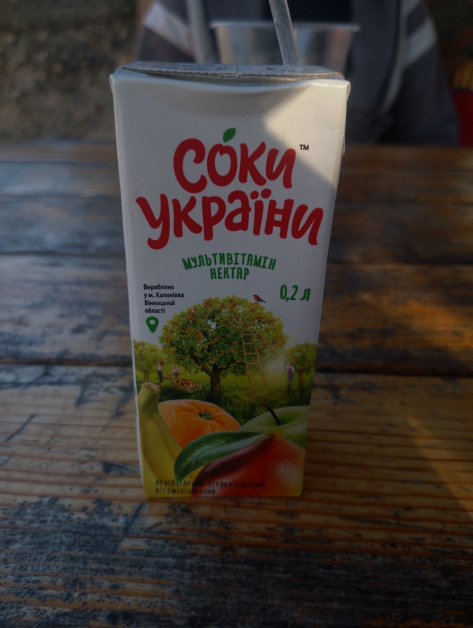 A juice box sits on a wooden table. The juice box is white with red text that reads "СОКИ УКРАЇНИ" which translates to "JUICES OF UKRAINE". Below that, it says "МУЛЬТИВІТАМІН НЕКТАР" meaning "MULTIVITAMIN NECTAR". Further down, it states "Вироблено у м. Калинівка Вінницької області" which indicates it was produced in the city of Kalynivka, Vinnytsia Oblast, Ukraine. The image on the box depicts an orchard with a ladder leaning against a tree. It also shows various fruits like an orange, banana, and apple. The box contains 0.2 liters of juice. The wooden table appears weathered, with visible grain and some scratches. The background is blurred but suggests an outdoor setting with dappled sunlight and possibly people. The overall scene is a close-up shot of the juice box, highlighting its branding and contents. The location specified on the juice box is Kalynivka, Vinnytsia Oblast, Ukraine, not Ustyvytsia.