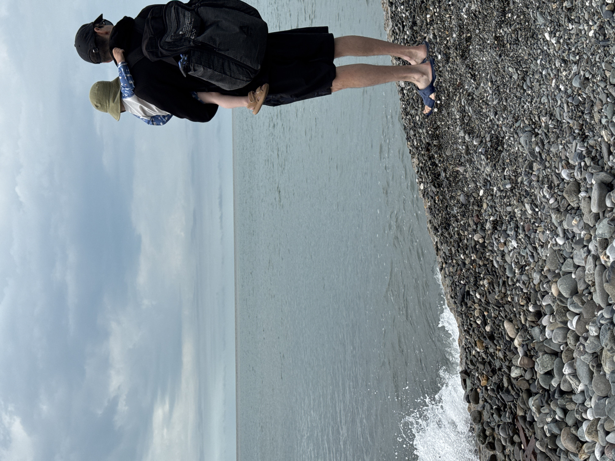 A father wearing a black cap, black backpack, and black shorts carries a child wearing a green sun hat on his back while standing on a pebble beach. The father wears blue sandals, and the child wears tan sandals. The sea borders the pebble beach, with the sky above. The location is Sochi, Russia.