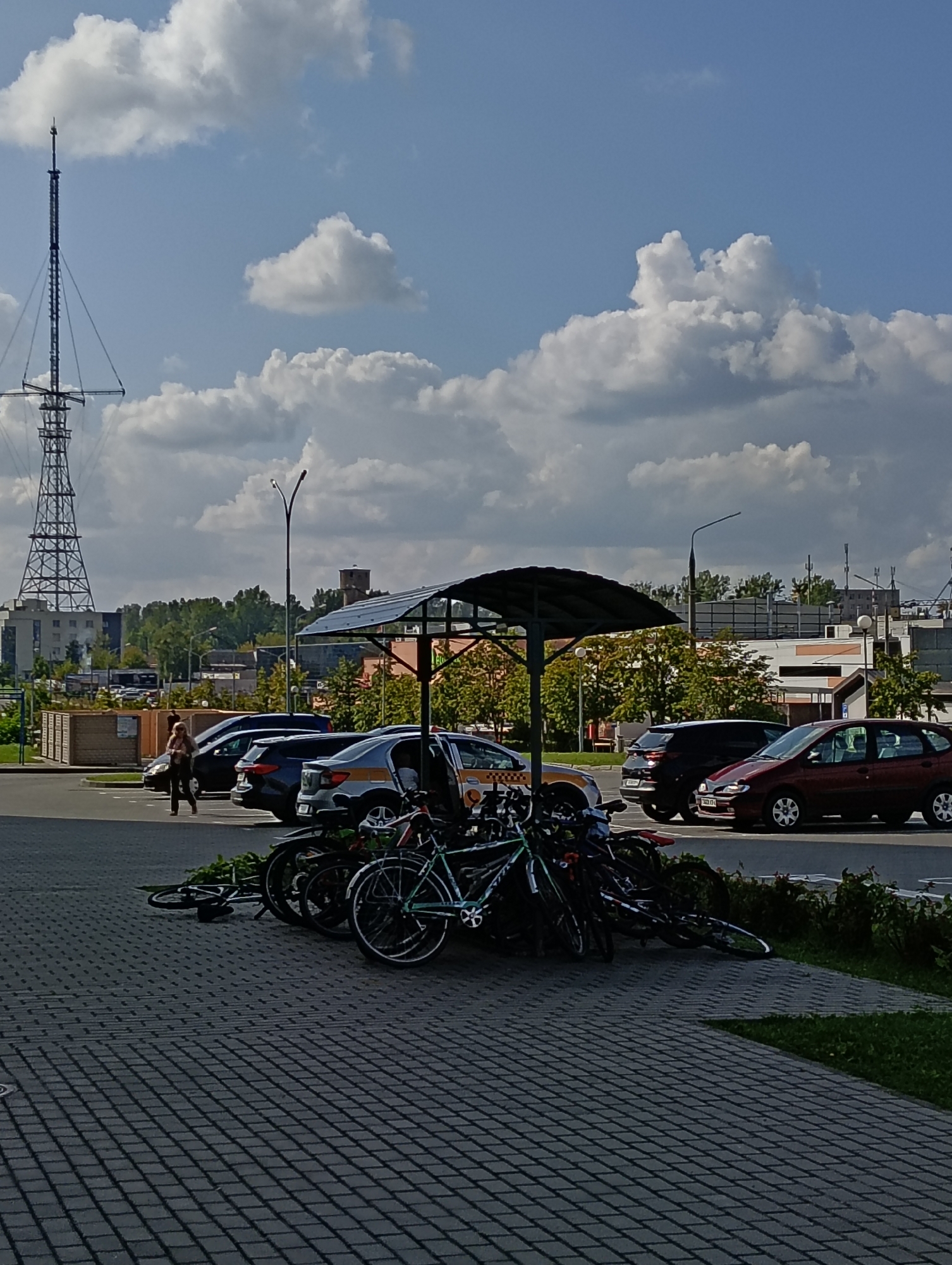 A street scene in Hrodna, Belarus features a parking lot with several cars and bicycles. A tall radio tower is visible in the background. A woman is walking across the parking lot. There is a covered area with solar panels over a row of bicycles. The sky is blue with white clouds.