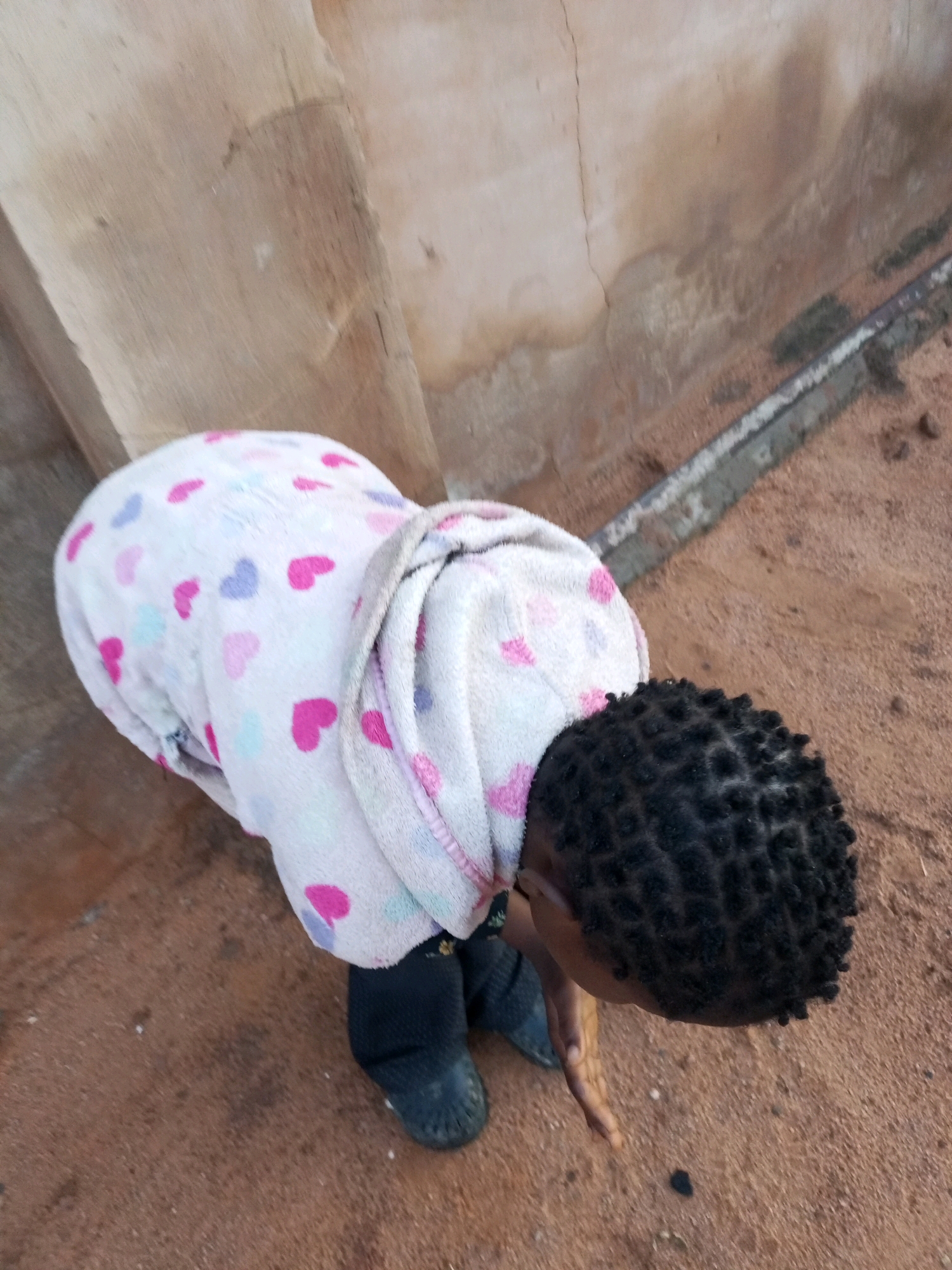 A child wearing a white fleece robe adorned with pink and purple hearts is bent over on sandy ground. The child has dark, braided hair. The background features a textured, light-colored wall with some cracks and discoloration. A dark line of cement or concrete runs along the ground, partially embedded in the sand. The scene appears to be outdoors in a rural or developing urban environment, possibly in a city like Tempure, Nigeria.