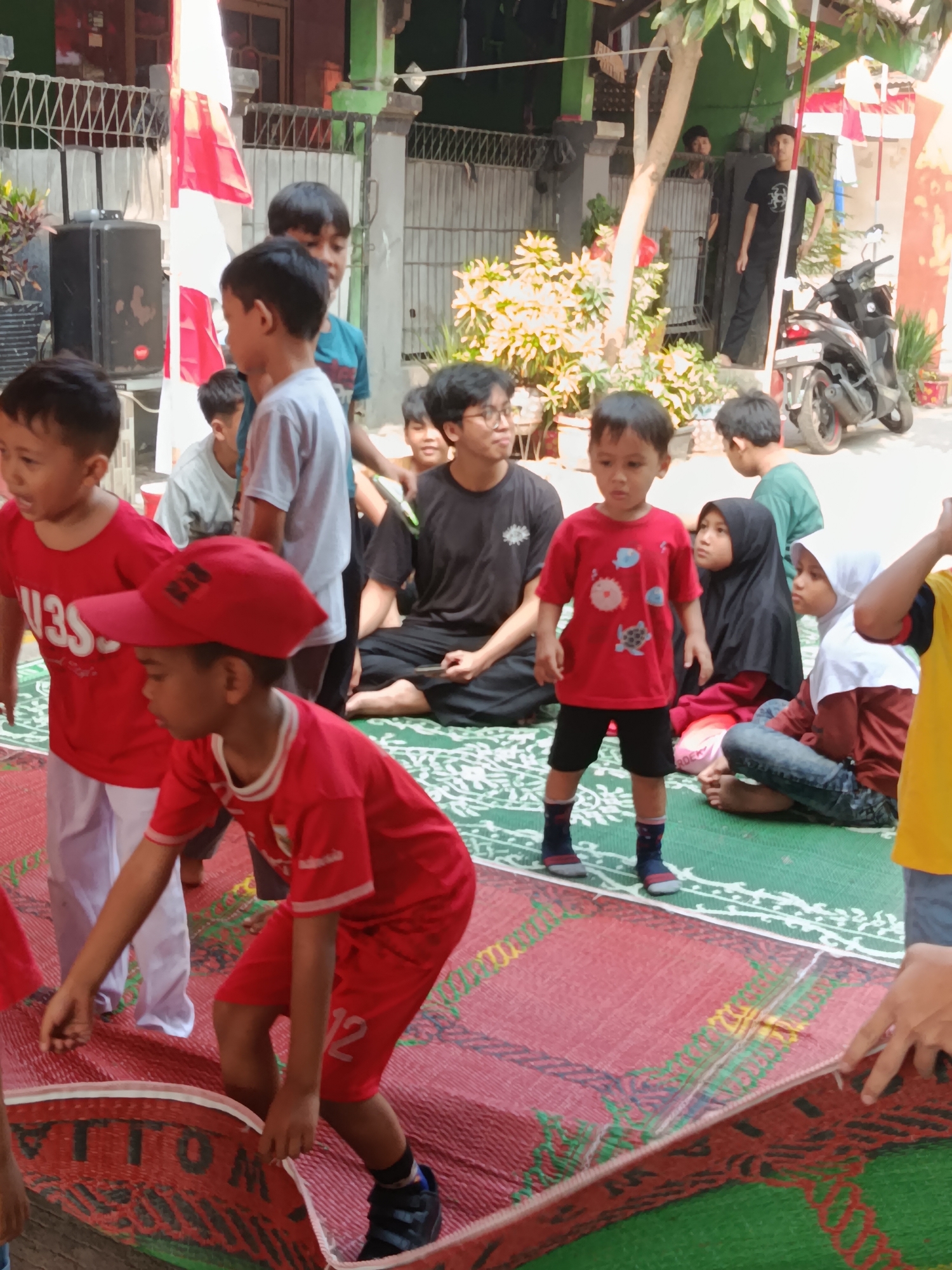 The image depicts an outdoor gathering in Wanasari, Indonesia, likely a community event or celebration given the presence of the Indonesian flag. In the foreground, several young children are the primary subjects. A boy wearing a red shirt and a red baseball cap with a white logo is bent over, appearing to adjust a rolled-up red and green mat. To his left, another young boy in a red shirt with the word "US" visible, is looking to the side. Behind them, a group of older children and adults are seated on mats. One man in a black t-shirt and pants is seated, holding what appears to be a phone. Another person in a black hijab is also seated, looking forward. To the right, a motorcycle is parked, and several potted plants are visible, suggesting a residential or communal outdoor space. The background features buildings with green walls and a white fence, and red and white Indonesian flags are prominently displayed on poles. The ground is covered with green and red mats, indicative of an event taking place on a soft surface. The overall atmosphere appears casual and family-oriented.