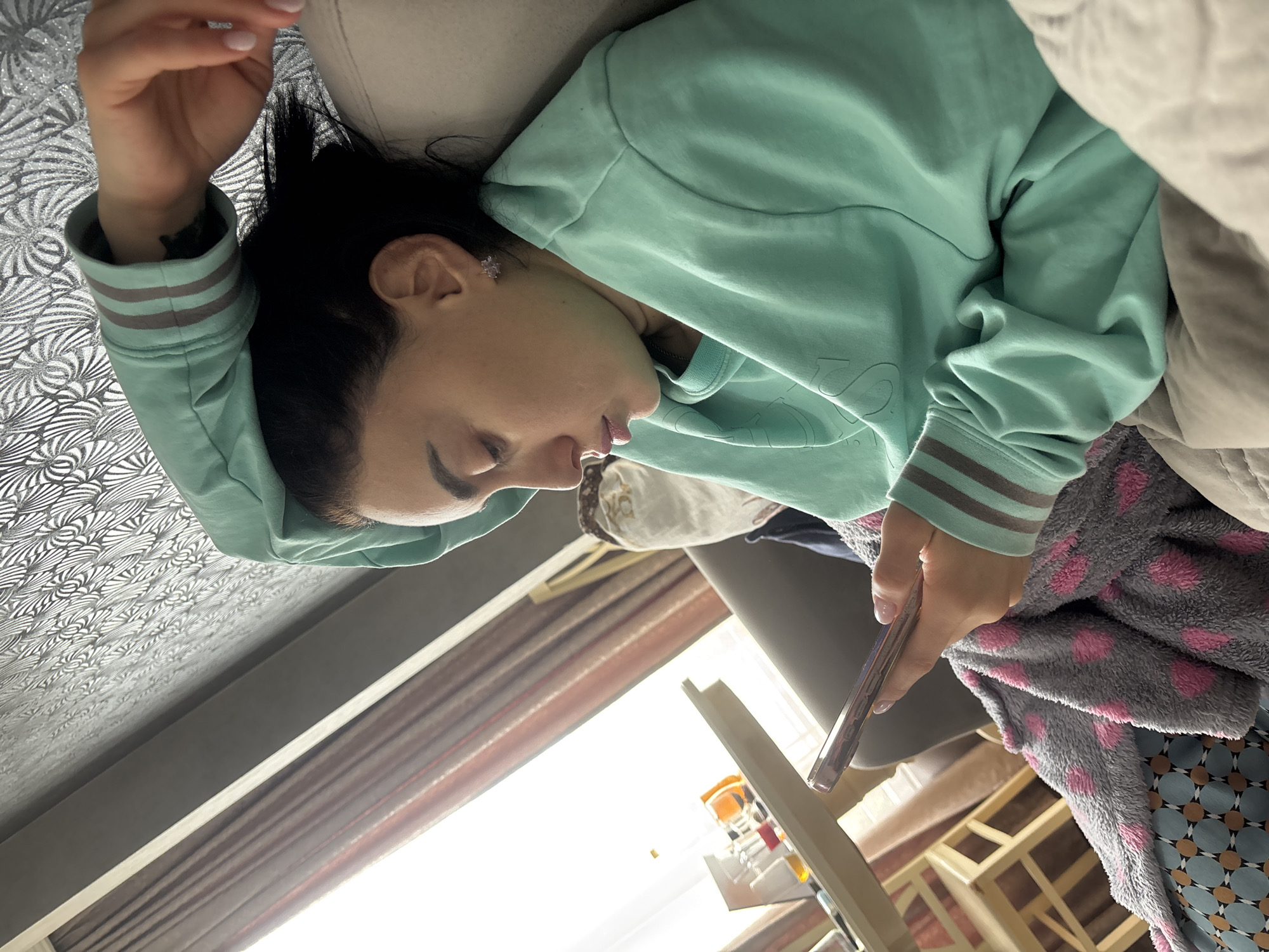 The image, originally rotated 90 degrees counter-clockwise, shows a young woman relaxing indoors, seemingly on a sofa or bed, engrossed in her smartphone. She has dark hair and wears a light aqua or mint green long-sleeved top featuring distinctive light blue and greyish striped cuffs. A small, sparkling, flower-shaped earring adorns her right ear, and her fingernails are painted a light pink or nude color.
She holds a smartphone with a pinkish-purple case in her right hand, her gaze directed towards its screen. A soft, grey blanket with pink polka dots is draped near her. In the background, a decorative white and silver patterned wallpaper or fabric covers a wall or headboard. Bright natural light floods in from a window, partially obscured by brown-striped curtains, indicating it is daytime. Other domestic furniture elements, including what appears to be a wooden chair frame, are vaguely visible in the sunlit area. The top she wears has embossed or embroidered text, but it is indistinct and illegible. The scene captures a private moment of leisure in a domestic setting, likely in Tbilisi, Georgia.