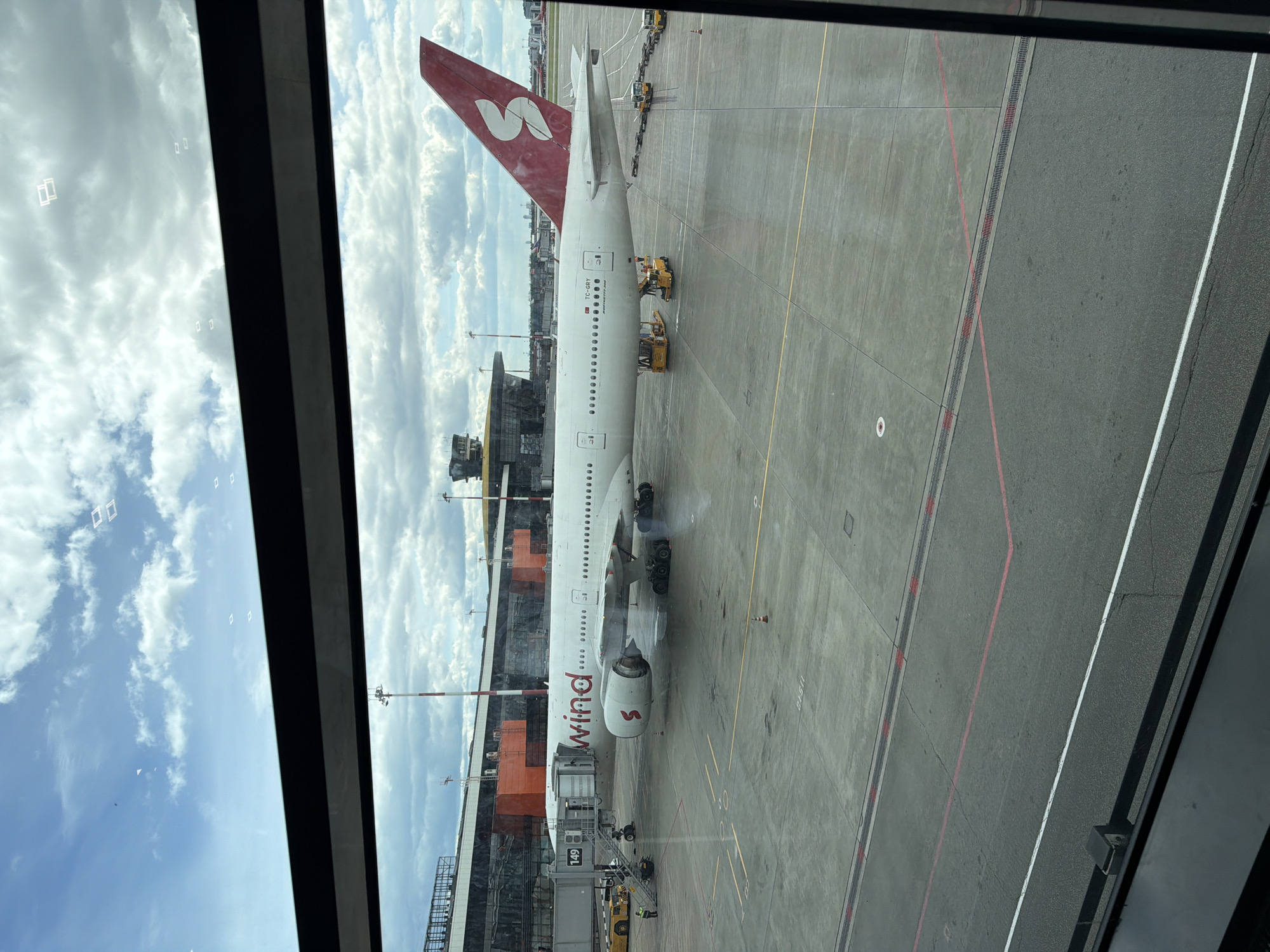The image shows a Nordwind Airlines Boeing 777-300ER aircraft, registration number TC-GRY, parked at an airport gate, viewed through a window. The aircraft is white with red accents on the tail, which features the Nordwind logo. The Nordwind logo is also visible near the front of the aircraft. The engine closest to the camera appears to be emitting exhaust. Several service vehicles are positioned around the aircraft. The jet bridge is connected to the aircraft's door. The airport's tarmac is gray with yellow and red markings. The sky is partially visible through the window, showing blue sky and clouds. Based on the airline and registration, the location is likely Sheremetyevo International Airport in Khimki, Russia.