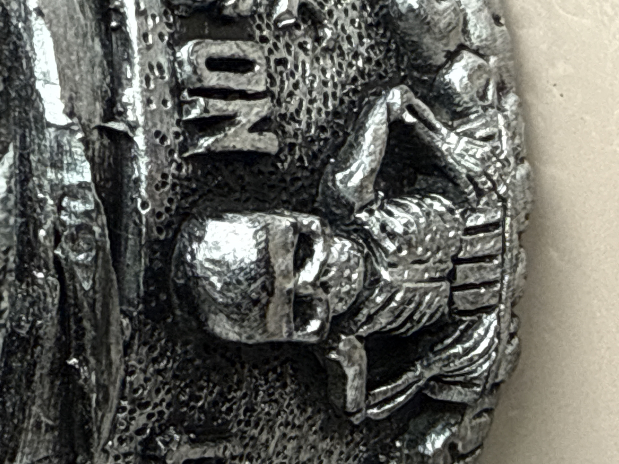 This image is a close-up, forensic shot of a dark, metallic sculpture. The sculpture depicts a skeleton sitting with its arms resting on its knees. The skeleton has pronounced ribs and a skull with empty eye sockets and a grin. Behind the skeleton, the texture suggests a rough, possibly rocky or pitted surface. To the right of the skeleton, the letters "NO" are clearly visible, embossed in the same metallic material. The lighting creates strong highlights and shadows, emphasizing the three-dimensional details of the sculpture. The background is out of focus and appears to be a neutral, possibly light-colored surface. No location in Tbilisi, Georgia, is visible or suggested by the image.