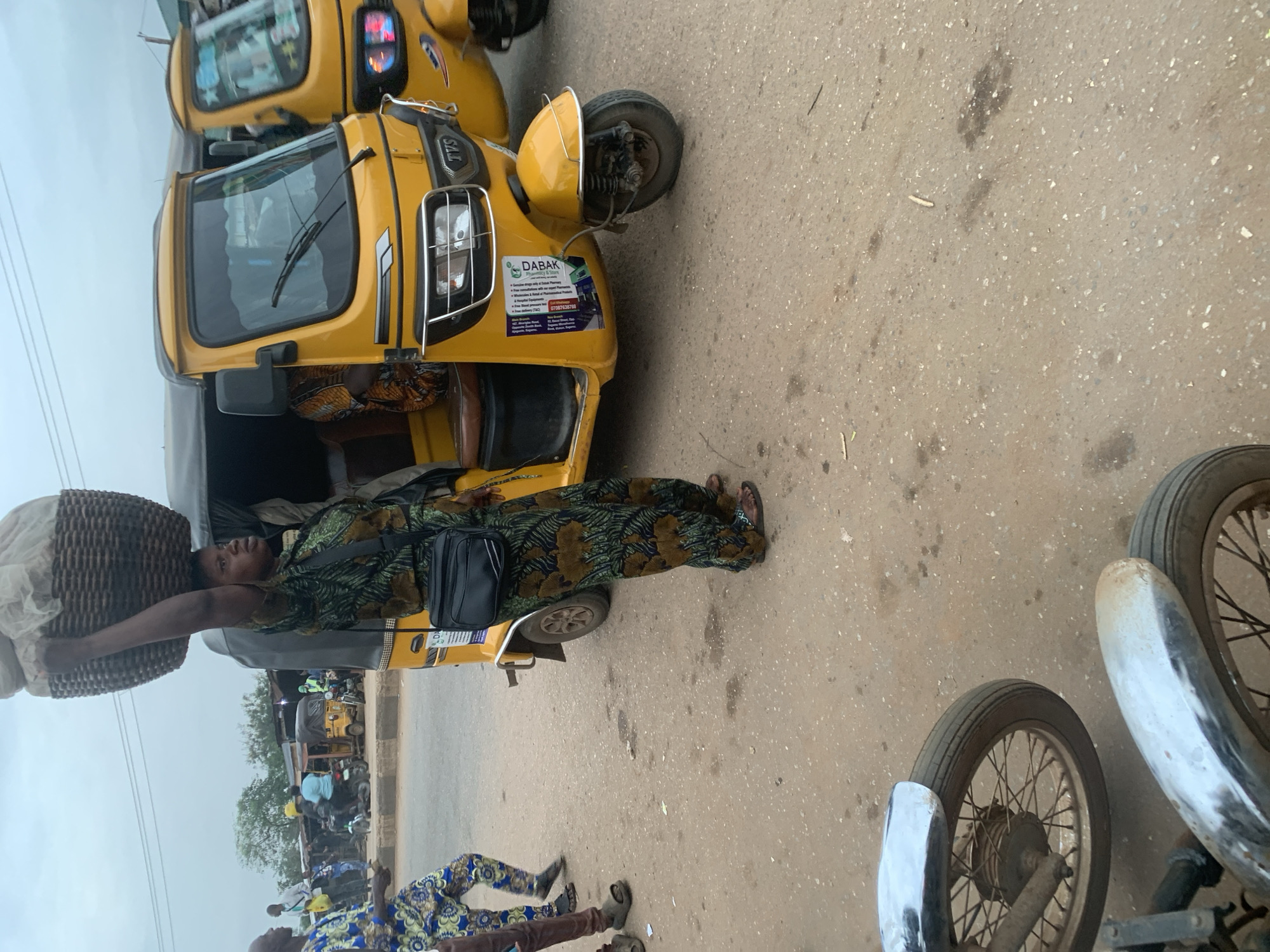 An overcast day in Shagamu, Nigeria, shows a woman balancing a large woven basket on her head while standing on a dusty road. She wears a long, green and brown patterned dress and a black satchel across her chest. Next to her is a yellow auto-rickshaw, with another identical vehicle visible behind it. The woman's right arm is raised, supporting the basket. In the background, other people and vehicles are present. The road surface is a mixture of dirt and asphalt, with some scattered debris. The scene suggests a busy marketplace or transportation hub.