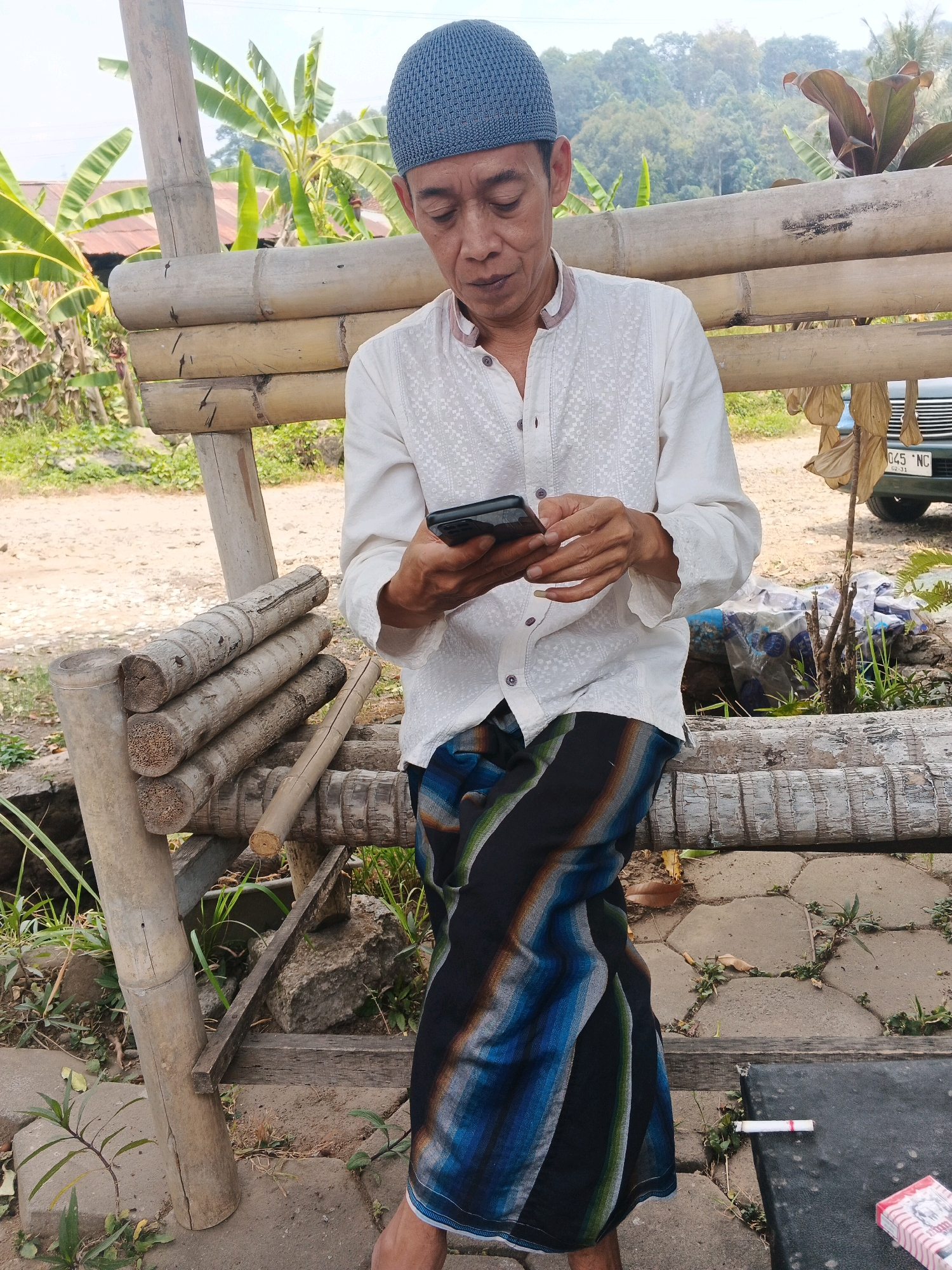The image depicts a middle-aged man, likely of Indonesian descent, sitting outdoors in what appears to be a rural or semi-rural setting. He is wearing a white, long-sleeved shirt with intricate embroidery and a blue, patterned sarong. On his head, he wears a blue skullcap. The man is holding a smartphone in his hands and appears to be focused on it.
He is seated on a rustic bench constructed from bamboo poles, with additional bamboo elements forming a backdrop. Behind him, lush green banana plants are visible, suggesting a tropical or subtropical climate. The ground is a mix of dirt and paving stones. In the background, a portion of a dark-colored vehicle is visible, with a license plate that reads "045 NC 02-11." The overall environment suggests a casual, outdoor gathering space, possibly in Ungaran, Indonesia, given the context. A single cigarette lies on a dark surface in the foreground, to the right of the man.