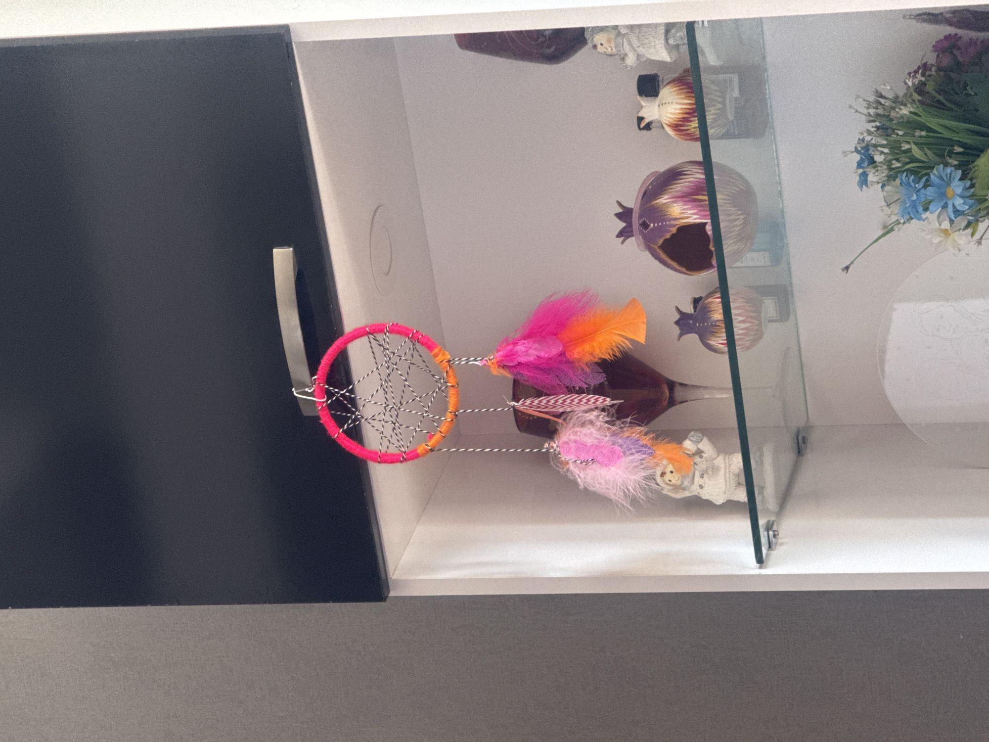 The image displays an interior shot of a decorative cabinet or shelving unit, showcasing various ornamental items. The main focal point is a colorful dreamcatcher with a vibrant pink and orange yarn-wrapped hoop, featuring a black and white string web design. Attached to it are strands of fluffy feathers in shades of bright pink, orange, and purple, some with striped patterns.
Inside the white-backed cabinet, behind a clear glass divider, are several decorative objects. On the upper shelf, partially visible, are small figurines. Below them, on a glass shelf, are two prominent, intricately patterned decorative pieces resembling pomegranates or stylized fruit, one in purple and gold tones, and another in brown and gold. A smaller, similar item is partly visible further back. On the bottom shelf, lying on its side, is a white figurine, possibly an angel or doll, with light, fluffy hair. To the right of the glass divider, on the top shelf, is a small bouquet of artificial flowers, primarily composed of delicate blue and white blossoms mixed with some purple flowers and green foliage.
To the left of the display cabinet, a dark grey or black cabinet door or panel with a modern silver handle is visible. Below the cabinet, a grey textured surface completes the immediate setting. The lighting suggests an indoor environment, possibly daylight or artificial light, illuminating the displayed items. No people are present, and no specific activity is taking place. There is no visible text. The image does not contain any visual cues to indicate the time of day, weather, or the specific location of Tbilisi, Georgia, provided in the context.