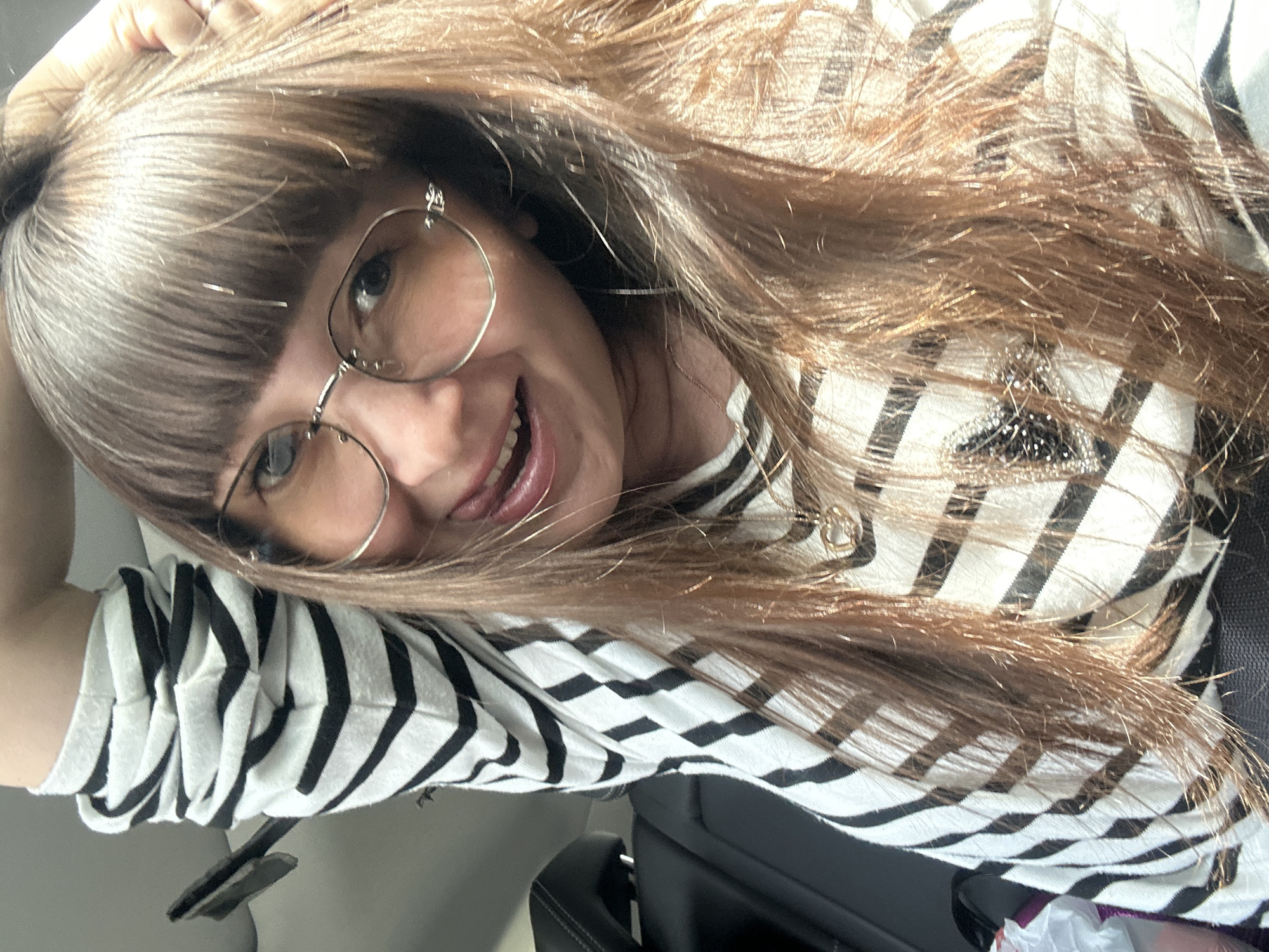 A woman with long, brown hair and bangs, wearing glasses and a striped shirt, is inside a vehicle. She is holding her hand up to her head and sticking her tongue out. The background shows a car interior with black seats. The image appears to be a selfie taken in the daytime. The location of Dokshitsa, Russia is not depicted in this image.