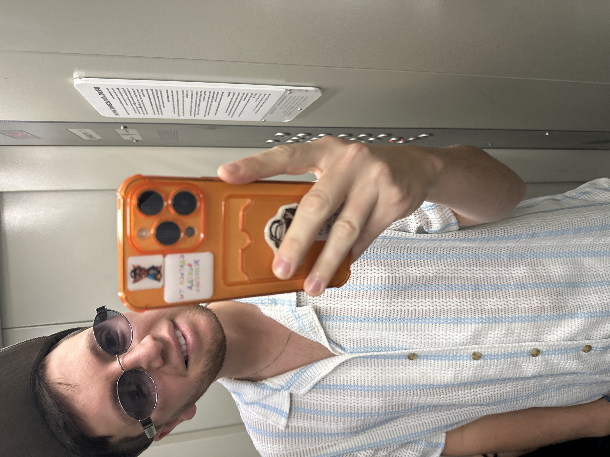 A young adult male is taking a selfie in an elevator. He is wearing a light-colored, short-sleeved shirt with vertical blue stripes, a gray baseball cap, and oval-shaped sunglasses. He is holding an orange-cased smartphone, which is reflecting the scene. On the phone case, there are two stickers: one with a cartoon character wearing a hat and a blue shirt, and another with colorful text. The elevator walls are gray metal, and a panel with buttons and a sign with text is visible on the right. The image is well-lit, with the focus on the man and his phone. The location is identified as Mirny, Russia.