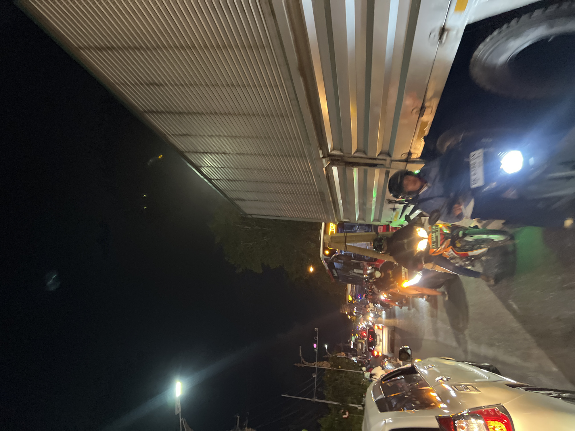 The image depicts a nighttime traffic jam in Denpasar, Indonesia. The scene is characterized by heavy traffic congestion, with numerous vehicles including cars and motorcycles filling the road. Streetlights illuminate the scene, casting long shadows and highlighting the headlights and taillights of the vehicles. A large truck dominates the right side of the frame, its metallic surface reflecting the lights. On the left, a white car is visible. In the center, several motorcycles with riders wearing helmets are part of the traffic flow. The overall atmosphere suggests a busy urban environment experiencing typical nighttime traffic challenges.