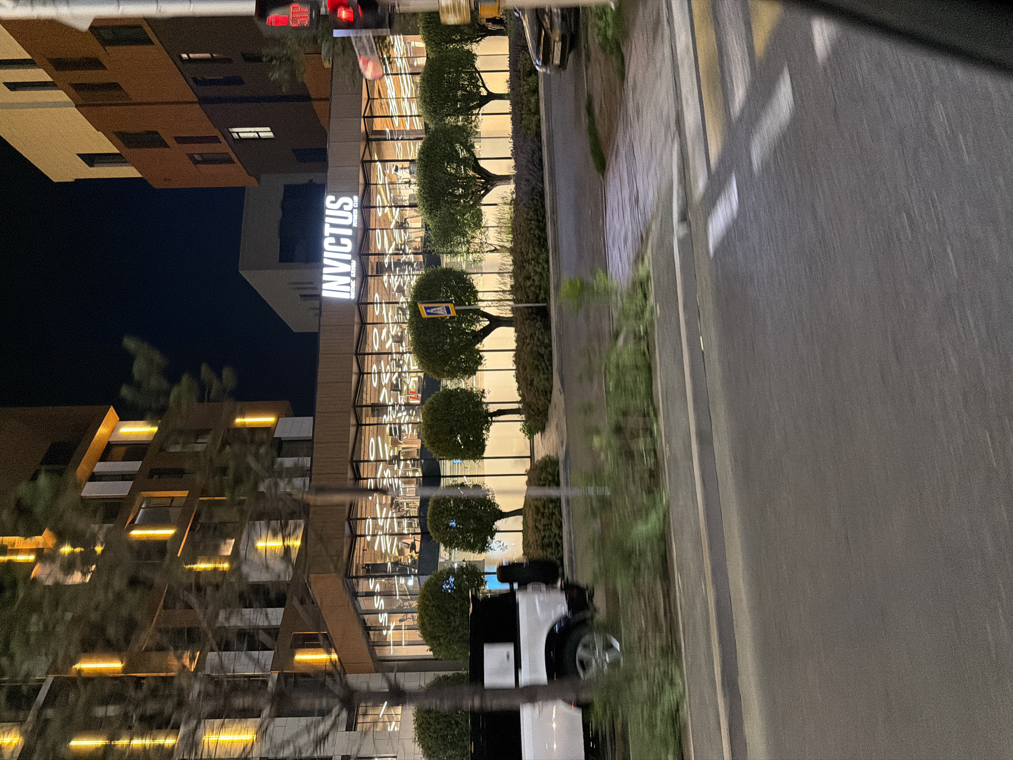 The image captures an outdoor night scene in Astana, Kazakhstan, featuring the "INVITUS FITNESS CLUB" building. The building has a modern architectural design with large glass windows, behind which gym equipment is visible, illuminated by warm interior lighting. The exterior is adorned with vertical golden lights. In front of the building, neatly trimmed spherical trees are planted in a row. A white SUV is parked to the left, partially obscured by foliage. The foreground shows a street with a pedestrian crossing marked by white and yellow stripes. Traffic lights are visible on the right, indicating a red light. The overall ambiance is one of urban nighttime activity.