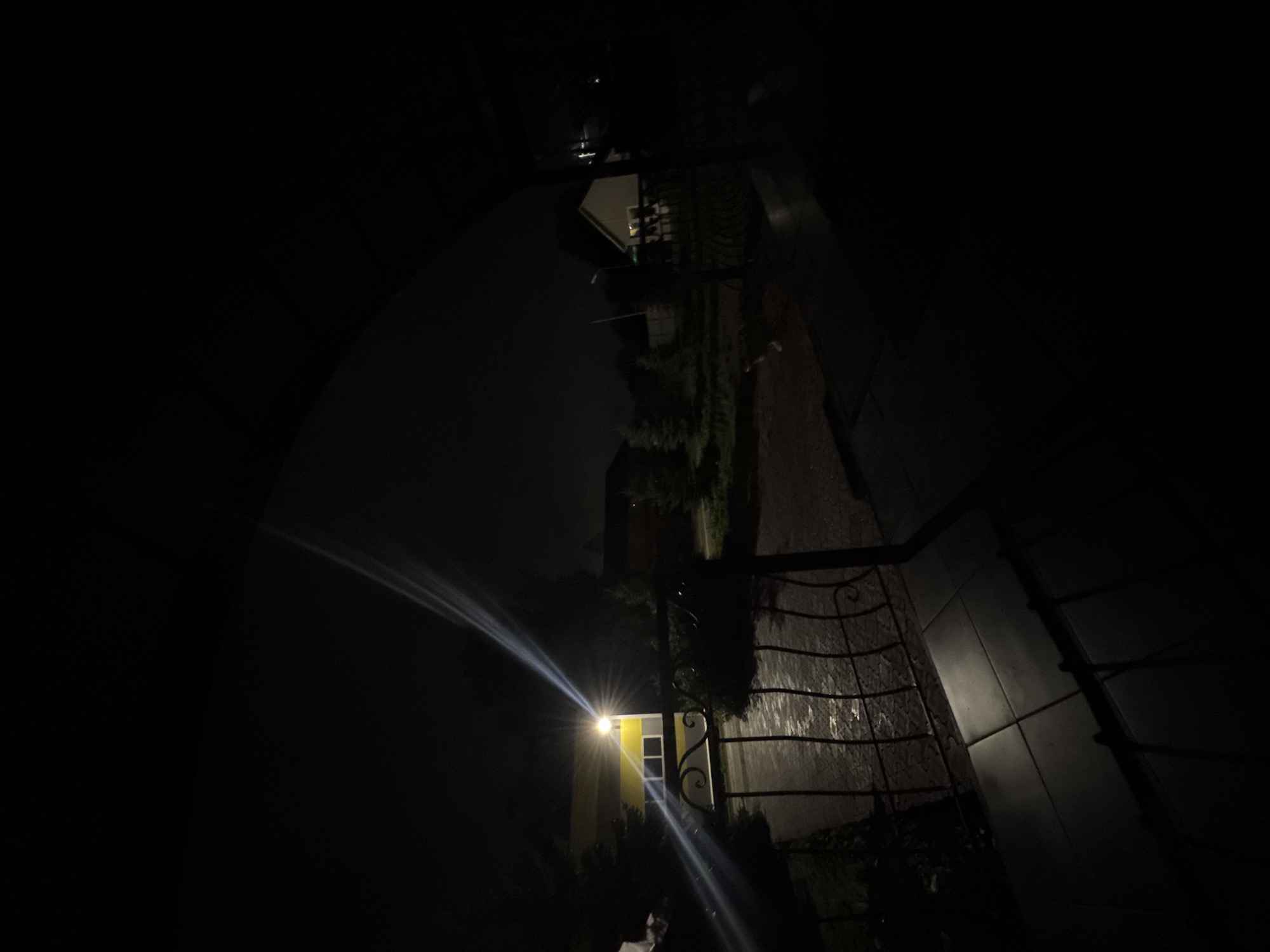 The image depicts a nighttime scene of a street in an unknown city. The foreground is dominated by a dark, wrought-iron gate and a tiled path casting long shadows. To the left, a brightly lit yellow building with white windows is visible, with a strong light source emanating from it, creating a lens flare effect across the image. Beyond the gate, a wet, paved area reflects the light. In the mid-ground, several trees and two houses are visible. One house, to the right, is illuminated by an interior light. The sky is dark and overcast, suggesting a lack of ambient light or a cloudy night. The overall atmosphere is dark and slightly eerie, with the intense light from the building being the primary focal point.