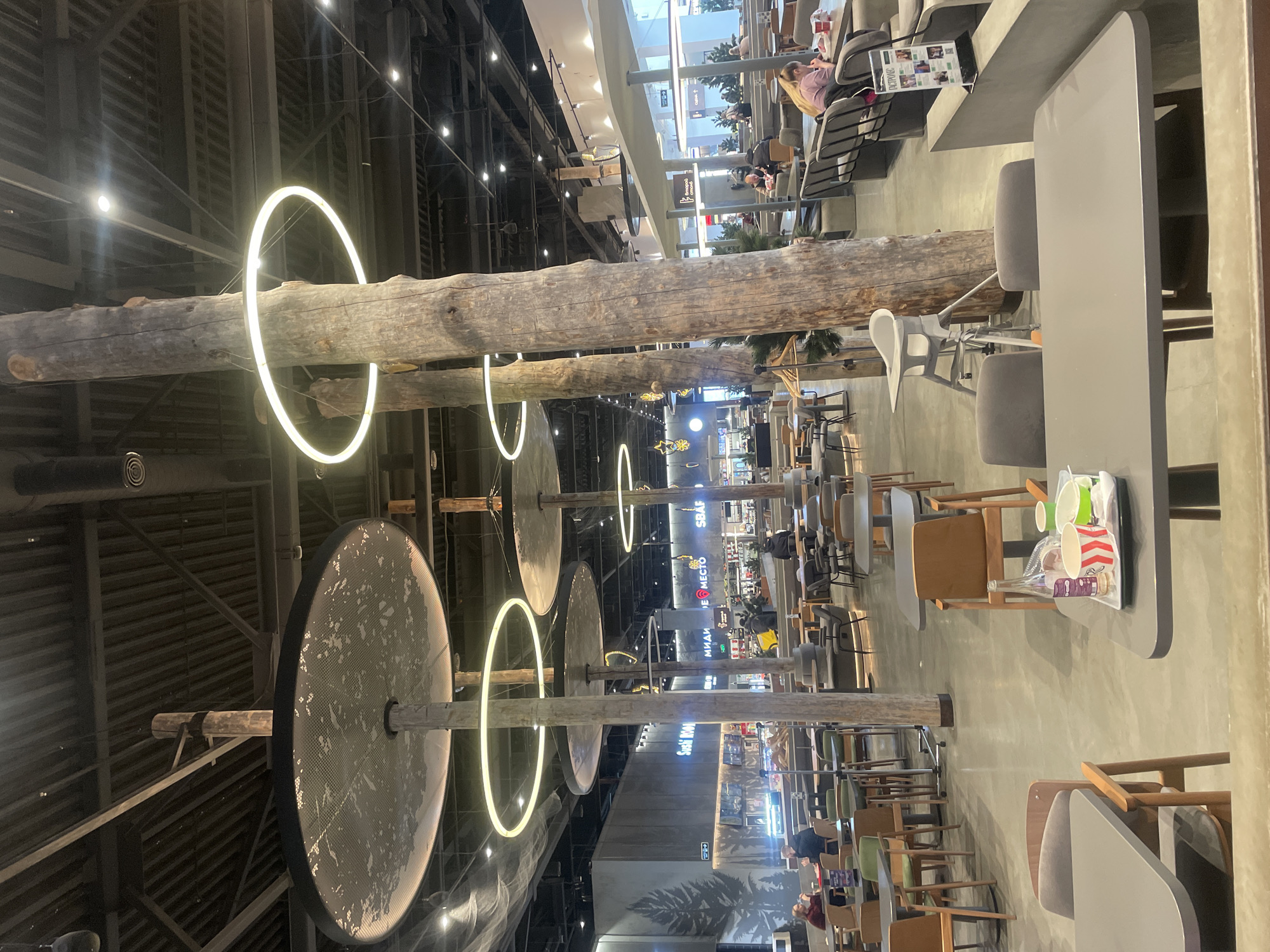 This image depicts a vibrant indoor public space, likely a food court or a large cafe within a modern shopping center, located in Krasnoyarsk, Russia. The setting is characterized by a blend of rustic and contemporary design elements.
The most striking features are several massive, rough-hewn wooden logs that span horizontally across the ceiling area, serving as both structural and decorative elements. Attached to these logs are bright, glowing circular LED lights, creating a unique and warm ambiance. Below these logs, large, dark, circular panels, possibly acoustic or lighting fixtures, are suspended from additional wooden posts. The ceiling above features exposed industrial-style girders and discreet spot lighting.
Numerous tables and chairs are arranged throughout the space, suggesting a casual dining or lounge area. The furniture varies, with some tables being light grey and chairs featuring grey upholstery, while others are made of wood. Several people are visible, seated at these tables, some appearing to be eating or drinking, while others are engaged in conversation or resting. In the foreground, a tray on a table holds disposable cups and other remnants, indicating recent use.
In the background, the space opens up further, revealing more seating areas and what appear to be commercial establishments. Elements like glass railings and escalators hint at a multi-level shopping complex. Visible text on signs includes "FOODPARK" and "Sushi BOM," along with other Cyrillic script such as "МИДИИ" (Mussels), suggesting diverse food options. The overall atmosphere appears relaxed and inviting, illuminated by a mix of natural light from above (partially visible through windows/skylights) and the distinctive artificial lighting from the circular fixtures.
