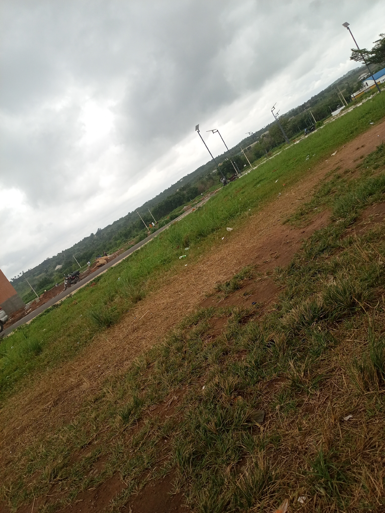 This is an oblique, medium shot of an outdoor scene in Iseyin, Nigeria, under a cloudy sky. The foreground is dominated by dry, brown grass and patches of reddish-brown dirt. A dirt path runs diagonally through the grassy area. In the mid-ground, a paved road is visible, with a few motorcycles parked on the side and several people walking or standing near the road. Some construction appears to be underway, with piles of rocks and exposed earth. A few streetlights with solar panels are erected along the roadside. The background features a hilly, tree-covered landscape. On the far right, a white building with blue trim is partially visible. The overall impression is one of a developing area in a somewhat rural or suburban setting.