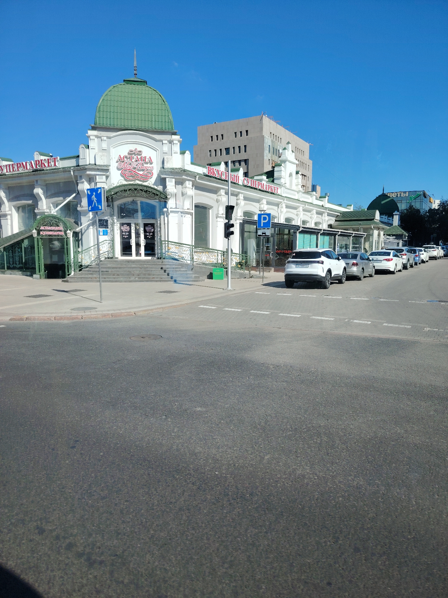 This is a daytime, eye-level, wide shot photograph taken from a street in Astana, Kazakhstan. The composition is dominated by a white, historic building with ornate architectural details, a green domed roof, and multiple entrances. The building appears to be a supermarket, as indicated by Russian signage that reads "АСТАНА" (Astana) and "ВКУСНЫЙ СУПЕРМАРКЕТ" (Delicious Supermarket).
In front of the building, there is a pedestrian crossing and a parking area where several cars are parked in a line. The cars are predominantly white and silver sedans and SUVs. A white SUV is parked closest to the camera, with its rear facing the viewer. A blue pedestrian crossing sign is visible on a pole near the building's entrance.
The sky is clear and blue. In the background, a modern, multi-story concrete building rises above the historic structure, suggesting a mix of old and new architecture in the urban environment. To the right, another building with a green domed roof and signage that includes the word "ЦВЕТЫ" (Flowers) is partially visible.
The foreground of the image consists of an asphalt road with white lane markings. The overall scene depicts a street view in a city, likely a commercial or mixed-use area, on a clear day. There are no people clearly visible in the photograph.