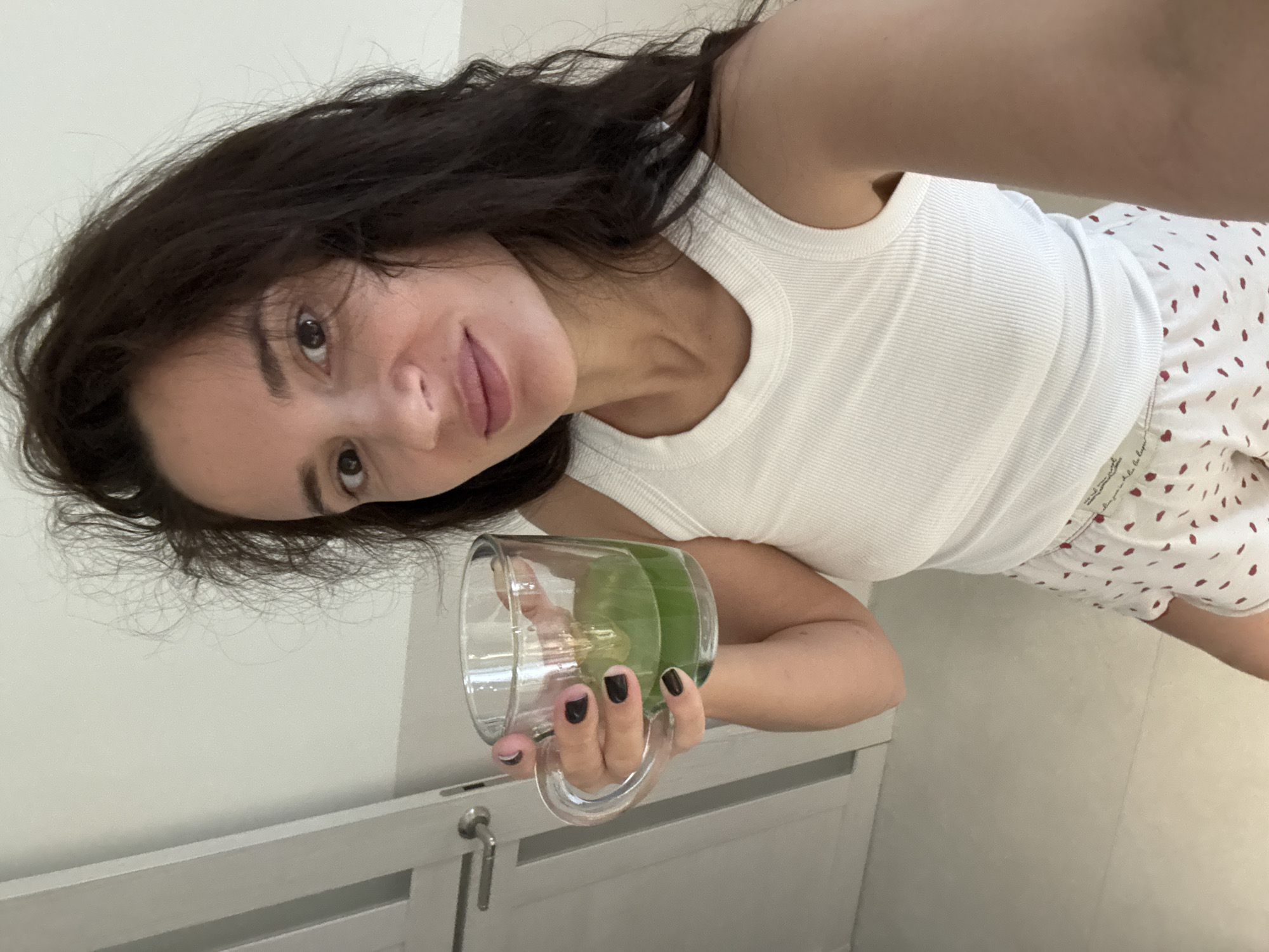 An adult female is depicted in a chest-up self-portrait indoors. She has long, dark brown, wavy hair, appearing somewhat disheveled, and dark eyes looking directly at the camera. Her complexion is light, and she has a subtle, closed-mouth smile. She is wearing a white ribbed tank top and white shorts with a red heart or dot pattern. In her right hand, she holds a clear glass mug containing a bright green liquid, approximately one-third full. Her fingernails are painted with a dark, possibly black, polish. The background consists of plain light-colored walls and a light-colored door or cabinet on the left side of the frame, featuring a silver-colored handle. The floor is also light-colored. The scene appears to be an interior space, possibly a residence. The location in the city of Chaikovsky, Russia, cannot be visually confirmed from the provided image content.