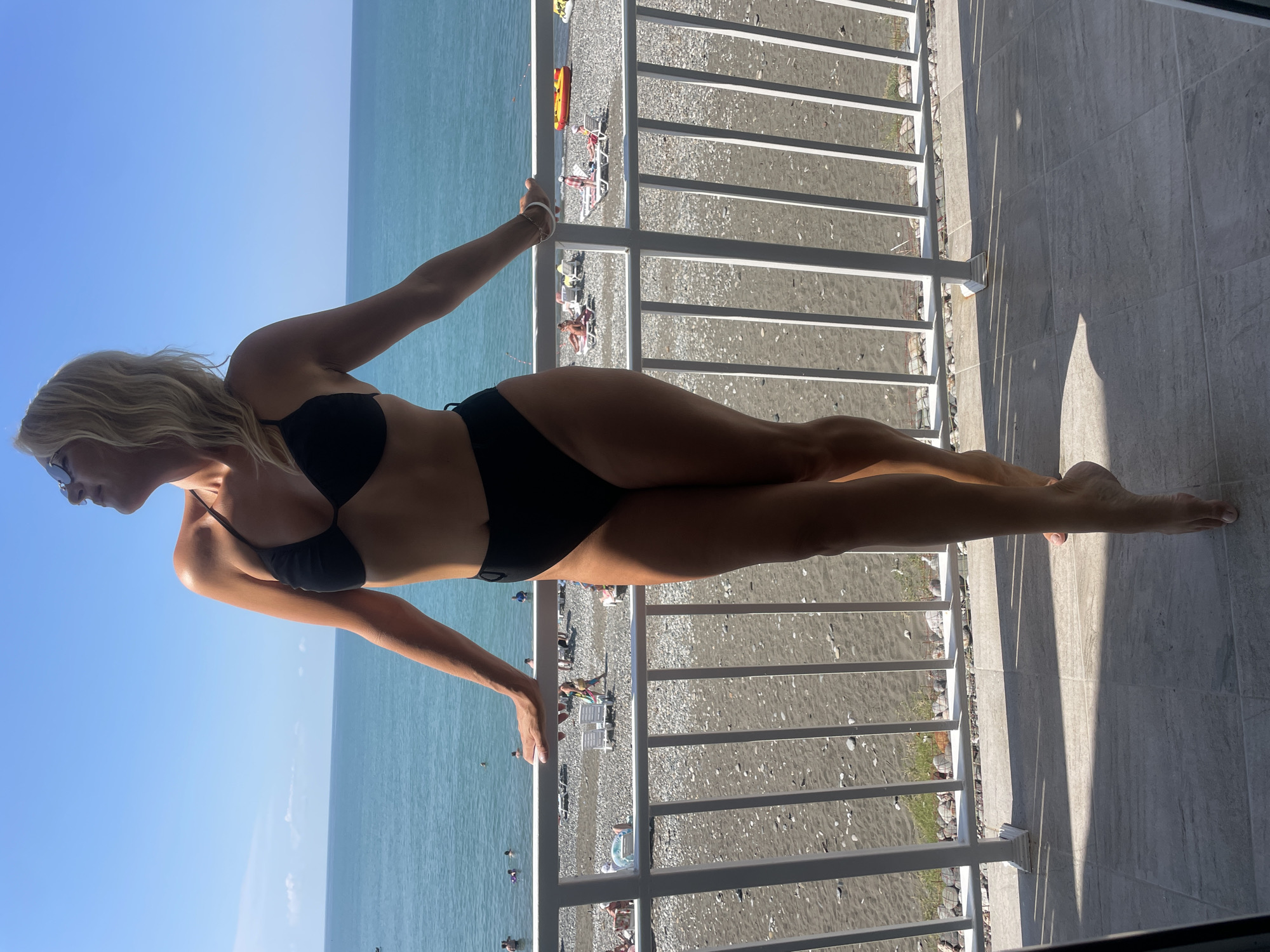 In the city of Gagra, Abkhazia, a woman stands on a balcony overlooking the sea. She is wearing a black bikini and sunglasses, with her head tilted back and eyes closed, basking in the sun. Her arms are resting on a white railing, and her body is turned slightly towards the camera. In the background, the turquoise water of the Black Sea stretches to the horizon under a clear blue sky. The beach below is covered with dark pebbles, and several people are visible lounging on chairs or wading in the water. The balcony floor is tiled, and shadows from the railing fall across it. The overall scene depicts a relaxed and sunny day at the seaside.