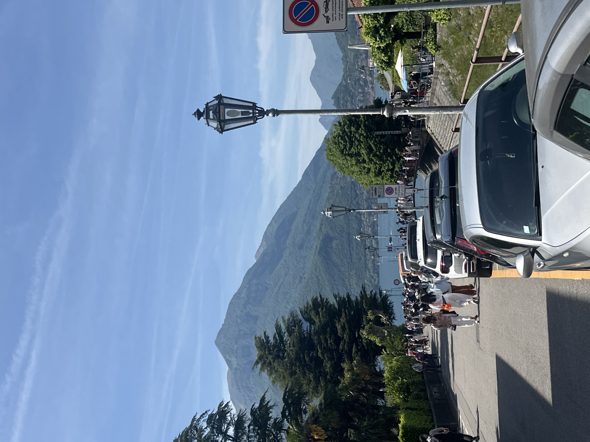 Captured from a street level perspective in the Italian town of Gittana, the image presents a landscape dominated by mountains, buildings, and a row of parked vehicles. A line of people is walking down the street to the left of the parked vehicles. The backdrop features a mountain range under a clear blue sky. Closer to the foreground, trees and a streetlamp with a decorative design are seen. A "No Parking" sign with a red circle and diagonal line is visible above the street lamp. The photo's orientation is vertical.