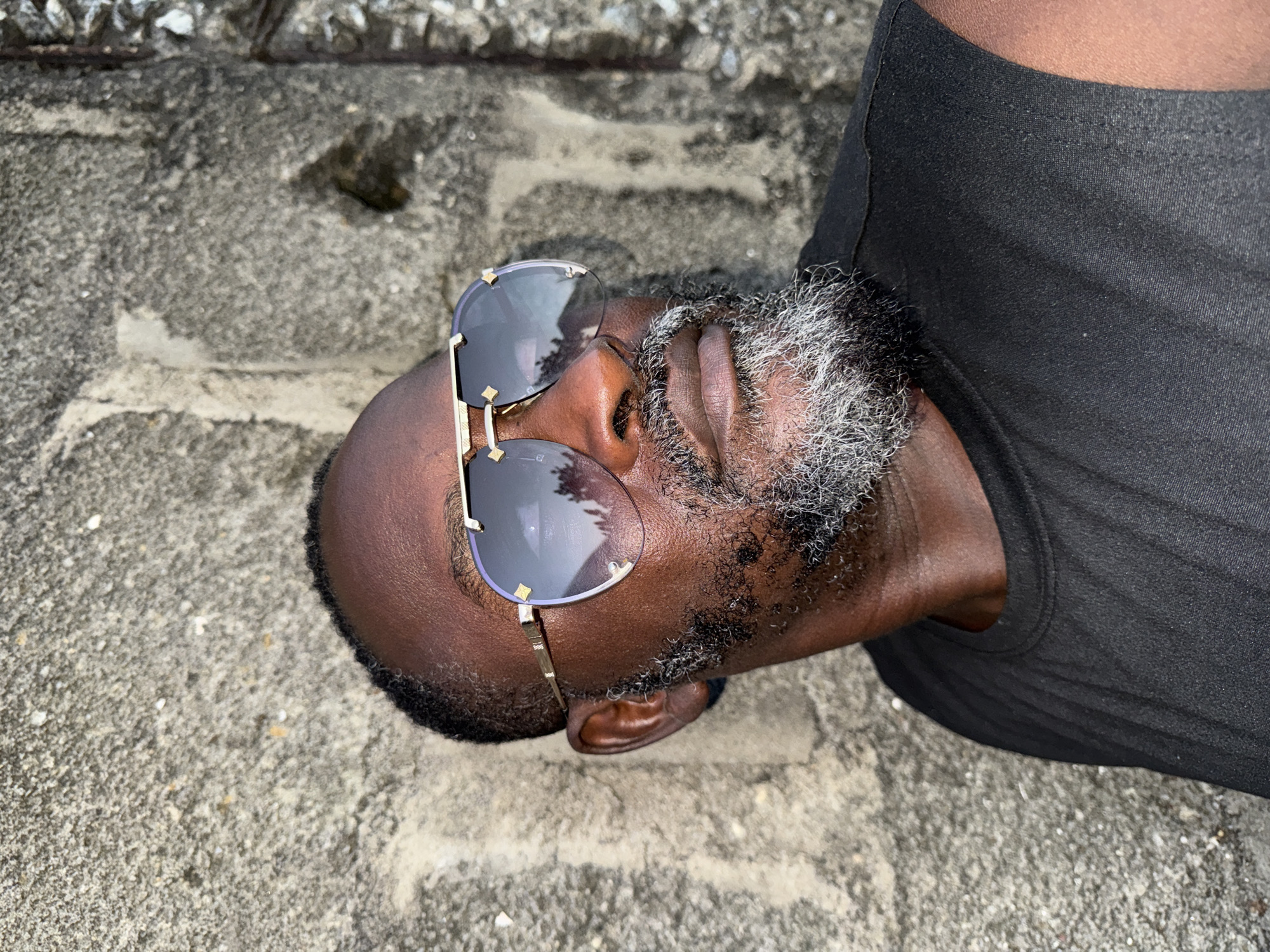 The image shows a close-up, overhead shot of a Black man wearing sunglasses and a black t-shirt. He has a salt-and-pepper beard and is lying on a concrete surface. The man's eyes are closed, and his head is tilted back, suggesting he might be resting or sleeping. The concrete surface appears textured and weathered, with some scattered pebbles. The lighting suggests it could be daytime, possibly overcast given the lack of strong shadows. The overall mood is one of repose. The setting is identified as Ado, Nigeria. There is no visible text in the image.