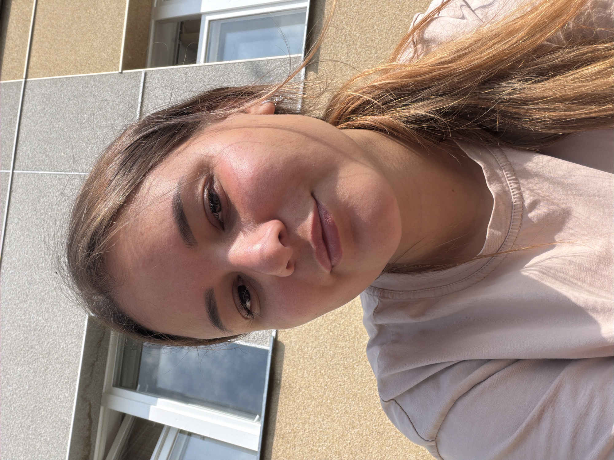 A young woman with light brown hair is posing for a selfie outdoors in front of a building. The building's facade is a textured beige color on the bottom, transitioning to a grey and then a tan color higher up. There are windows visible on the building, one with white trim and reflections of the sky. The woman is wearing a light-colored t-shirt. The lighting suggests it is daytime, with sunlight casting shadows. The woman is looking directly at the camera with a neutral expression. Her hair is pulled back into a ponytail, with some strands falling loosely around her face. She is wearing a small hoop earring in her visible ear. The image appears to have been taken at close range, focusing on the woman's face and upper body. The background suggests an urban environment, likely in Chaikovsky, Russia, given the context.