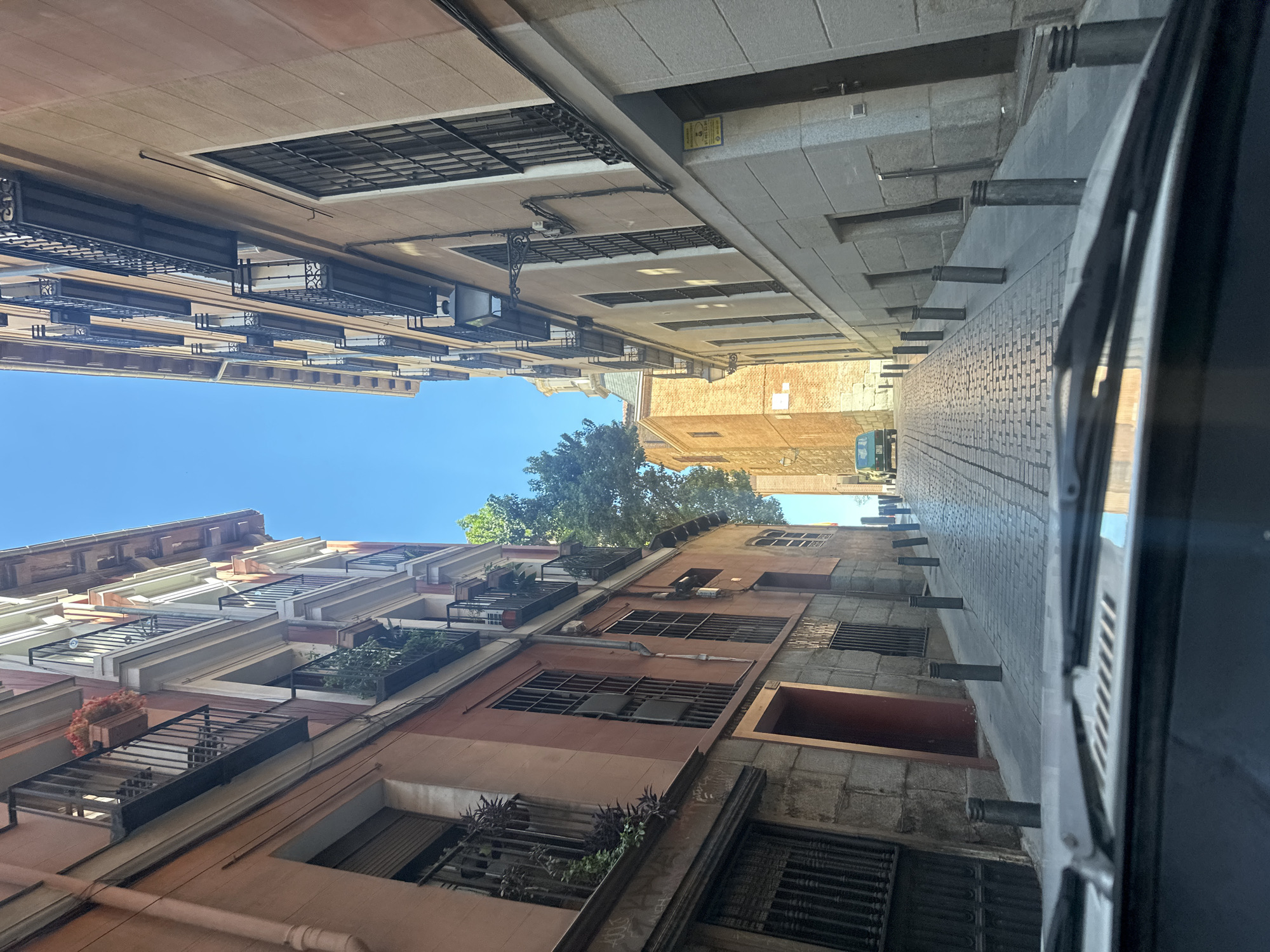 The image captures a view looking up a narrow urban street or alleyway, presumably from inside a moving vehicle, indicated by the dark car window frame visible on the right edge. The perspective is tilted, but when viewed upright, the street is flanked by multi-story buildings under a clear blue sky.
On the left side of the street, the buildings feature traditional European architecture with a mix of terracotta, beige, and white facades. Numerous balconies with intricate black wrought-iron railings extend from the windows, some adorned with potted plants and flowers, adding touches of greenery and color. Window grates are also visible on several levels.
The buildings on the right side of the street appear to have a more uniform grey or stone-like facade. Several small, dark cylindrical posts protrude horizontally from this building at regular intervals, possibly serving as architectural details or for street guiding. The pavement on this side of the street is made of light-colored, patterned tiles or bricks. A small yellow sign with illegible text is affixed to one of these buildings.
The bright sunlight suggests it is daytime, likely mid-day or afternoon, with strong shadows visible on the buildings and street. The sky is perfectly clear and blue, indicating good weather. In the distance, above the rooftops, the green canopy of trees can be seen. No people or animals are visible in the scene. The overall setting is a charming, historic urban environment, consistent with the context of Madrid, Spain.