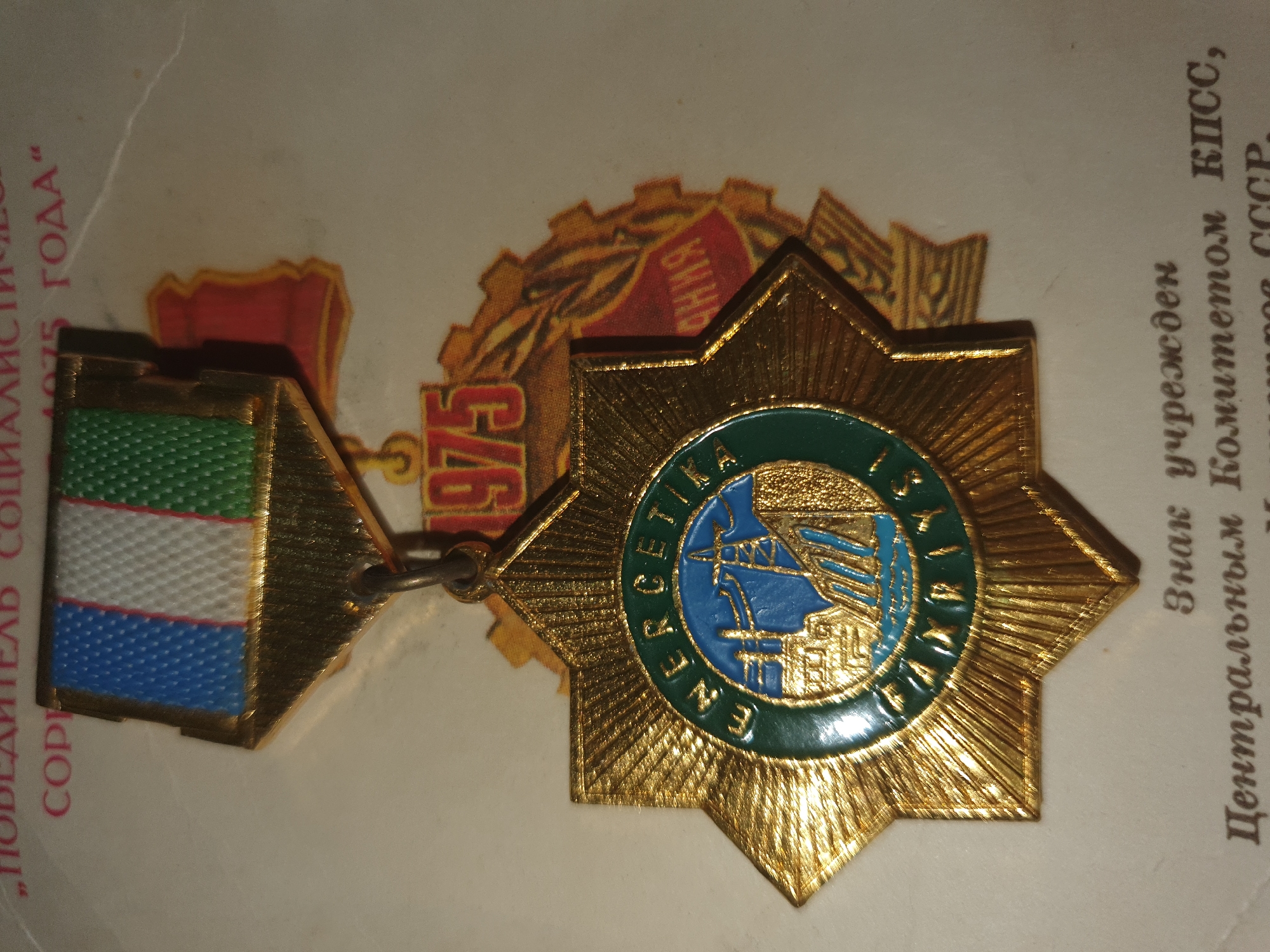 The image shows a close-up of a medal. The medal is gold-colored and features a star shape with radiating rays. In the center of the star is a circular emblem with a green border and a blue inner circle. The blue circle depicts a landscape with a power line tower, a dam, and buildings. Text in a circular pattern is visible around the inner blue circle. Above the star, a ribbon is attached, featuring vertical stripes of blue, white, and green. The ribbon is connected to a gold-colored backing. The medal appears to be resting on a light-colored surface with some text visible in the background, including the year "1975". There is no indication of the city of Tashkent, Uzbekistan, or any specific location within it. The image is a static, still life photograph of the medal.