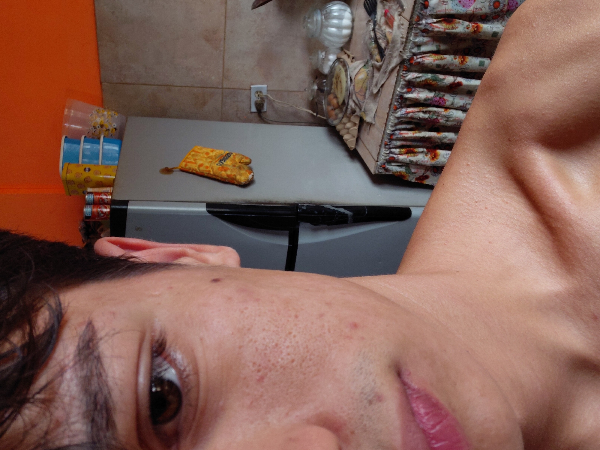 This image is a close-up, waist-up shot of a young person's right side of their face, neck, and shoulder, with a kitchen in the background. The person has dark hair, brown eyes, and visible pores and blemishes on their cheek. Their lips are slightly parted. The background shows a refrigerator with an oven mitt hanging on it, a shelf above the refrigerator with containers and cans, and a countertop with various items including an egg carton, a covered pan with food, and jars. The kitchen walls are tiled and painted orange. The location is identified as Santa Ana, Venezuela. The scene appears to be a domestic setting, likely a kitchen, during the day.