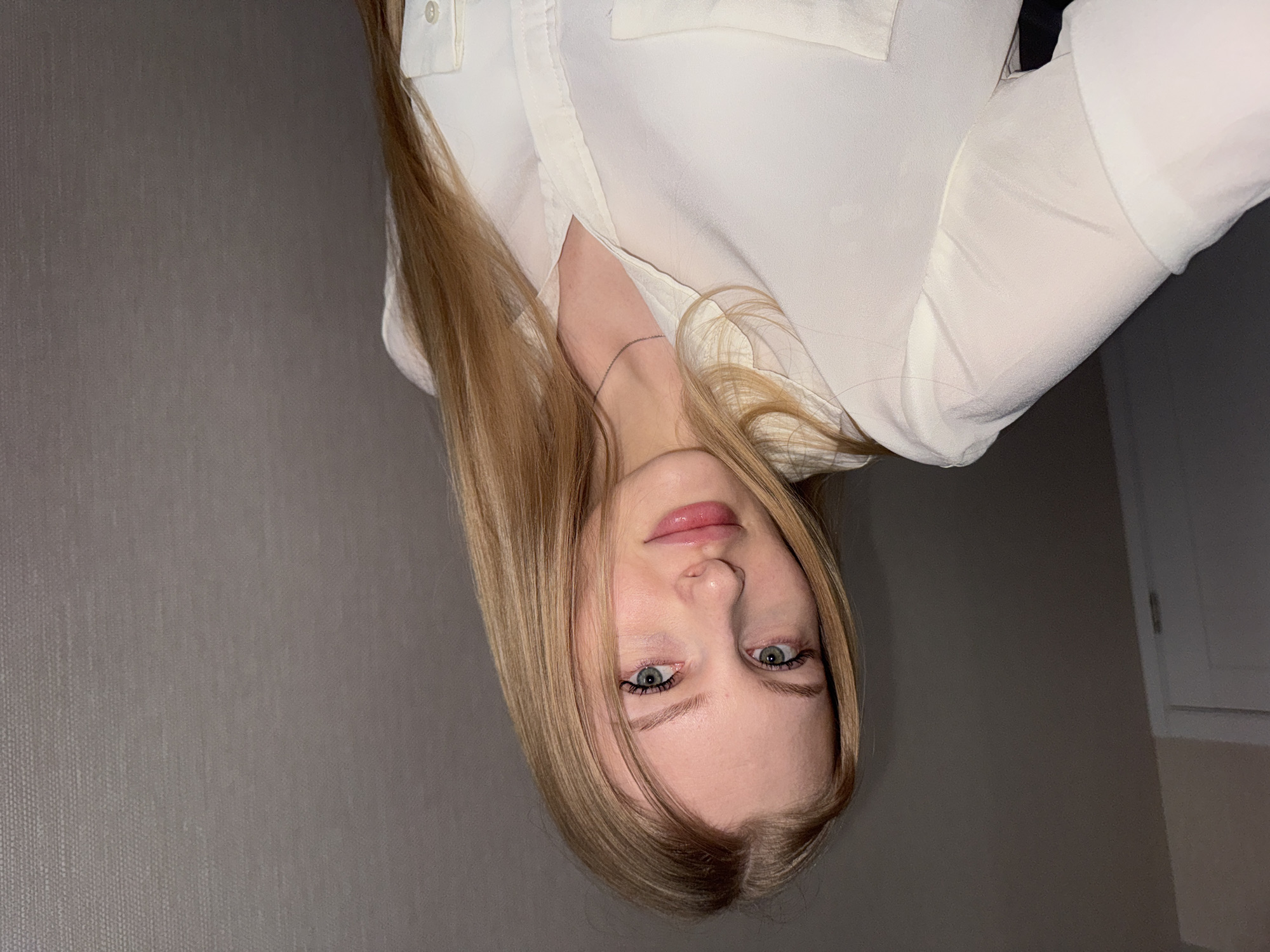 A portrait of a blonde woman with light eyes is shown in a rotated view. She is wearing a light-colored, buttoned shirt. The background consists of a gray wall and a glimpse of a white door or frame. The location is not specified, and there is no indication that the picture was taken in Perm, Russia.