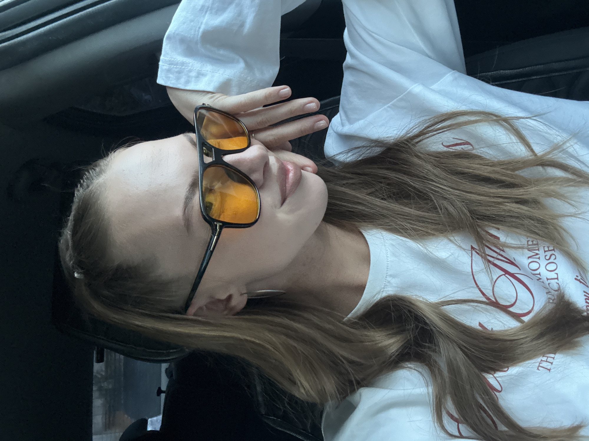 The media file shows a close-up, side-profile shot of a young woman wearing sunglasses and a white t-shirt. She is lying down, with her head resting against a dark surface, possibly a car seat. Her right hand is resting on her forehead, partially obscuring her face. She is wearing large, aviator-style sunglasses with orange lenses, which reflect the light. Her lips are slightly parted, and her eyes are closed or looking down, suggesting a relaxed or contemplative mood. Her long, light brown hair is spread out around her.
The white t-shirt has a graphic design on it, partially visible. The text reads, "MOMENT CLOSE" in a stylized font, with other letters also present but obscured by hair. The setting appears to be inside a vehicle, as indicated by the dark, textured surface behind her head, which resembles the interior of a car. The lighting suggests it is daytime. The overall impression is a casual and candid moment captured.