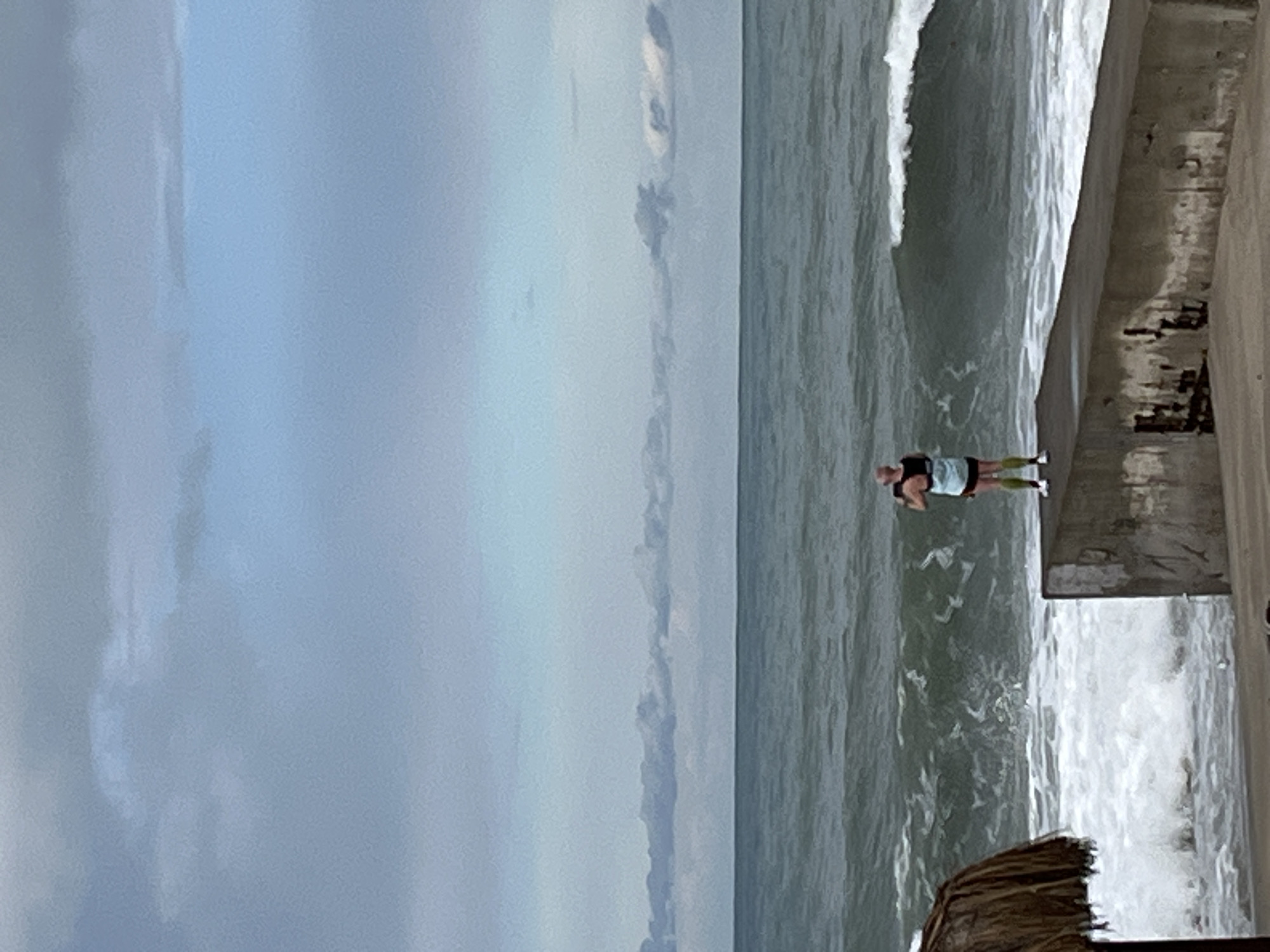 The image shows a person standing on the edge of a concrete structure next to a turbulent sea, possibly a beach or coastal area. The sky is overcast. The person is wearing athletic attire. The location is unknown.